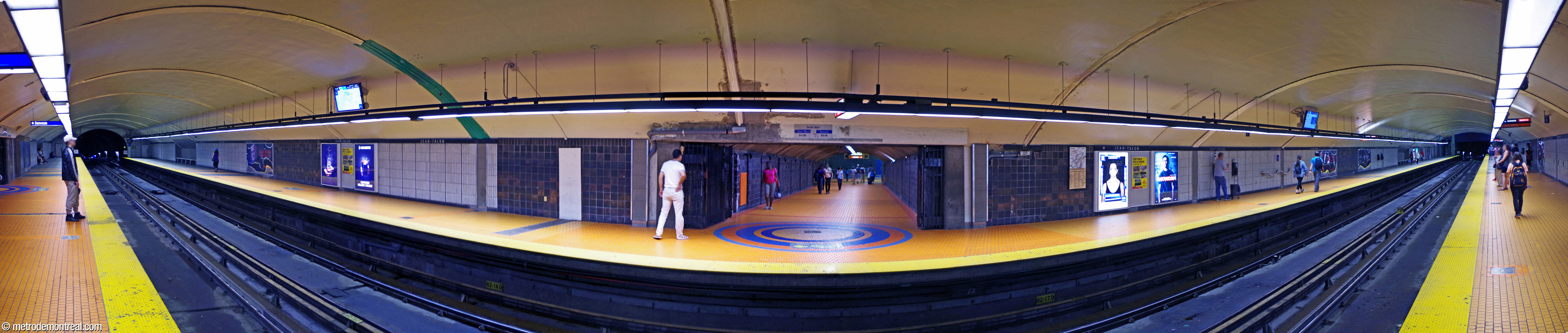 Panorama de la nef de la ligne orange, vue depuis le quai Côte-Vertu. La salle de contrôle est à droite. Au centre, un tunnel à partir du quai Montmorency mène vers la galerie est, offrant l'accès à la ligne bleue et à la sortie Saint-Hubert. Ce tunnel est marqué par un carrelage orange au sol avec une « rosace » bleue, des motifs créés par l'architecte de la station de la ligne bleue, Gilbert Sauvé.