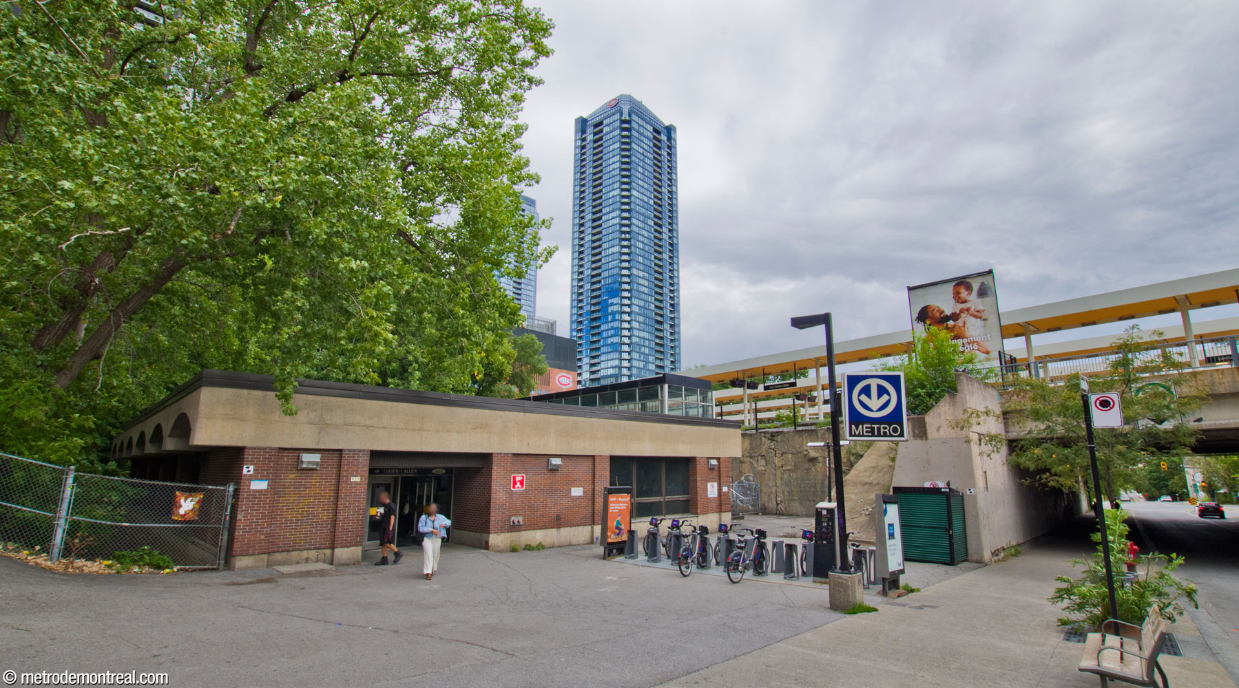 The station's unremarkable headhouse, located next to the abutment along the train station's platforms. Behind the headhouse is the enclosed passageway leading to the train station. The train station’s platform canopies are visible in the background.