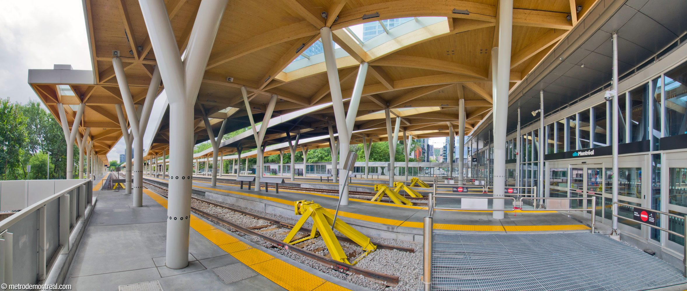Panorama of the platforms and terminal tracks serving the commuter train station, taken from platform 7/8. Each island platform serves two platform faces. The platforms are sheltered with long wood and glass canopies, supported on white Y-shaped pillars marked with the “o” from the Exo logo. At right, sliding doors lead into the station house. In the background, the connection between the commuter train and metro stations runs alongside the tracks; Rue de la Montagne is visible through the glass, showing that the ends of the tracks and platforms occupy a viaduct over the street.