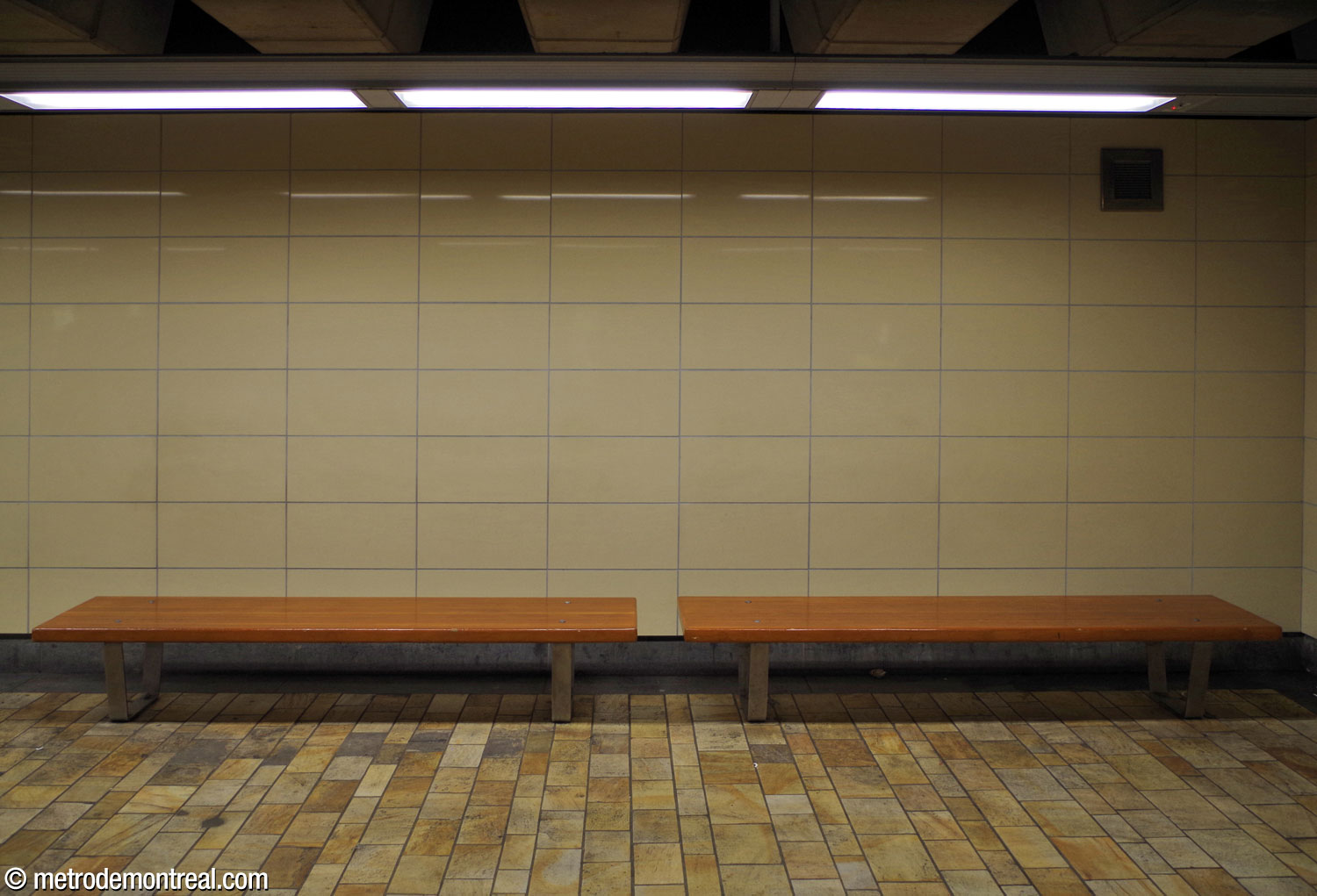 Two of the wooden benches on the departure platform, in front of a wall of beige tiles that do not match the cladding on most of the platform walls.