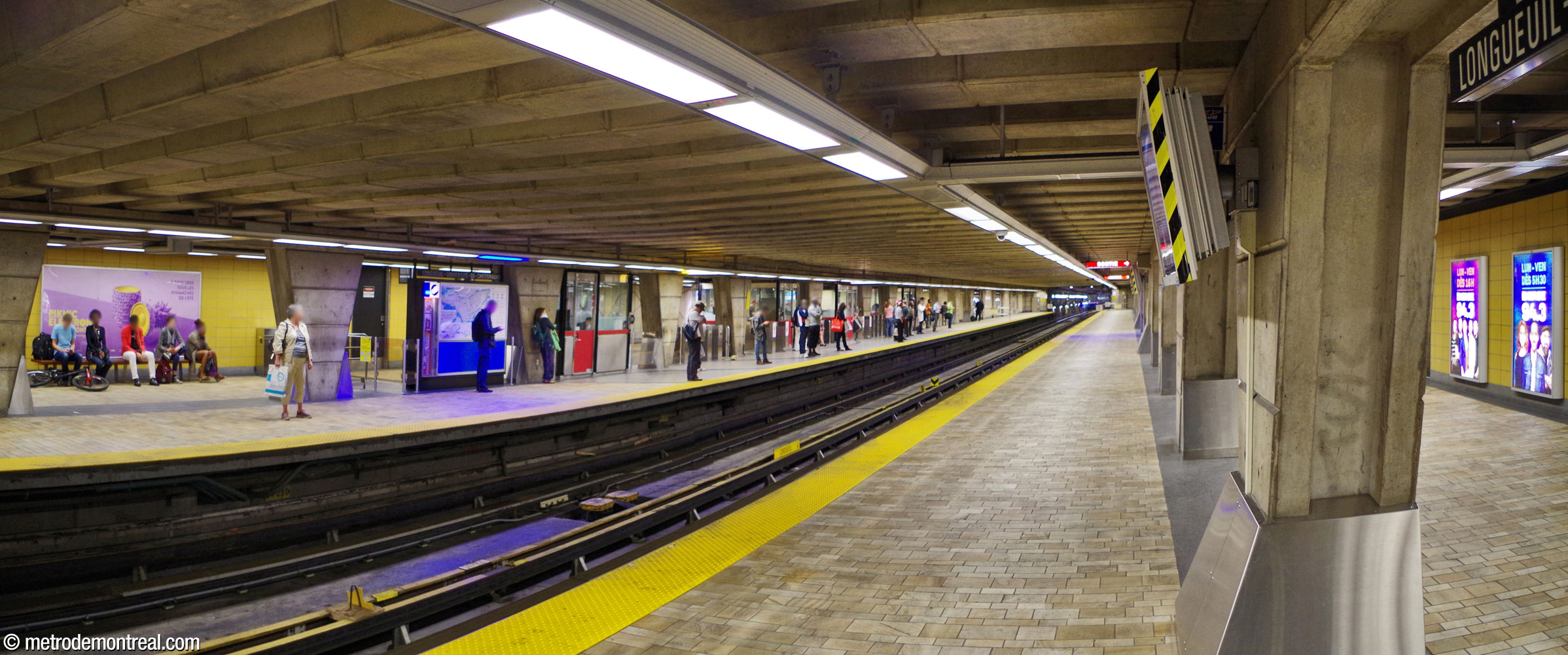 Panorama of the shallow trainroom, seen from the arrival platform. At left, passengers await a train on the departure platform; the turnstiles and booths of the ticket hall are behind them, amid the trapezoidal pillars.