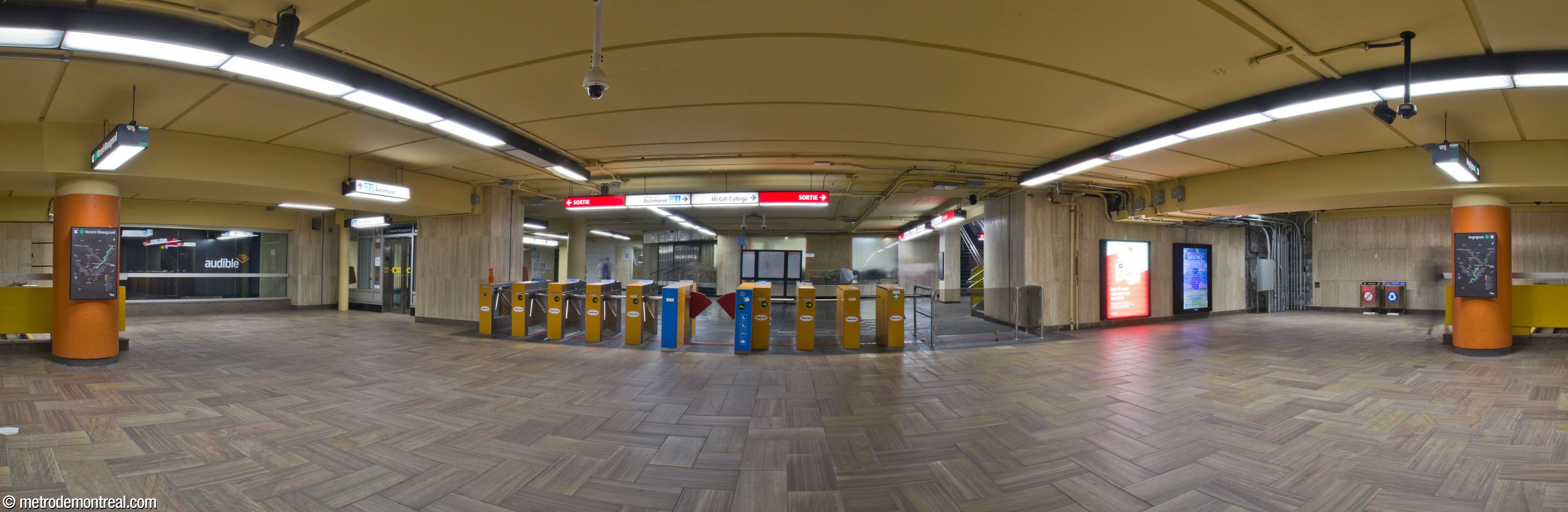 The fare-paid zone of the western ticket hall, with platform stairs on either side. The western concourse beyond the secondary gateline (centre) gives access to the McGill College, De Maisonneuve Ouest exits, to the western entrance to Centre Eaton, and to the REM, as well as to the elevator outside the fare-paid zone to the Angrignon platform and 705 De Maisonneuve Ouest headhouse. For its part, the elevator to the Honoré-Beaugrand platform is inside the fare-paid zone, at left.