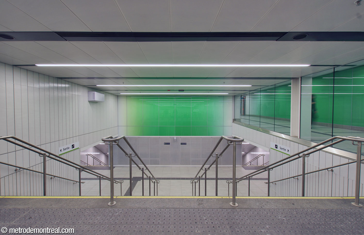 Inside the gateline of the north ticket hall, a broad staircase leads down to a landing, splitting into stairs down to the platforms. Above the landing is a decorative green gradient panel. At upper right, behind a glass wall, runs the corridor to the south ticket hall.