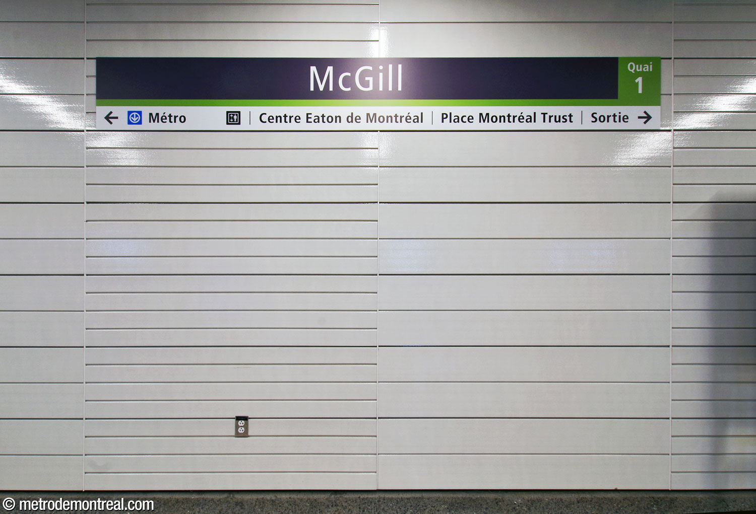 The platform walls, showing the station’s nameplate and its white enamelled metal wall plates of varying widths, placed horizontally at platform level.