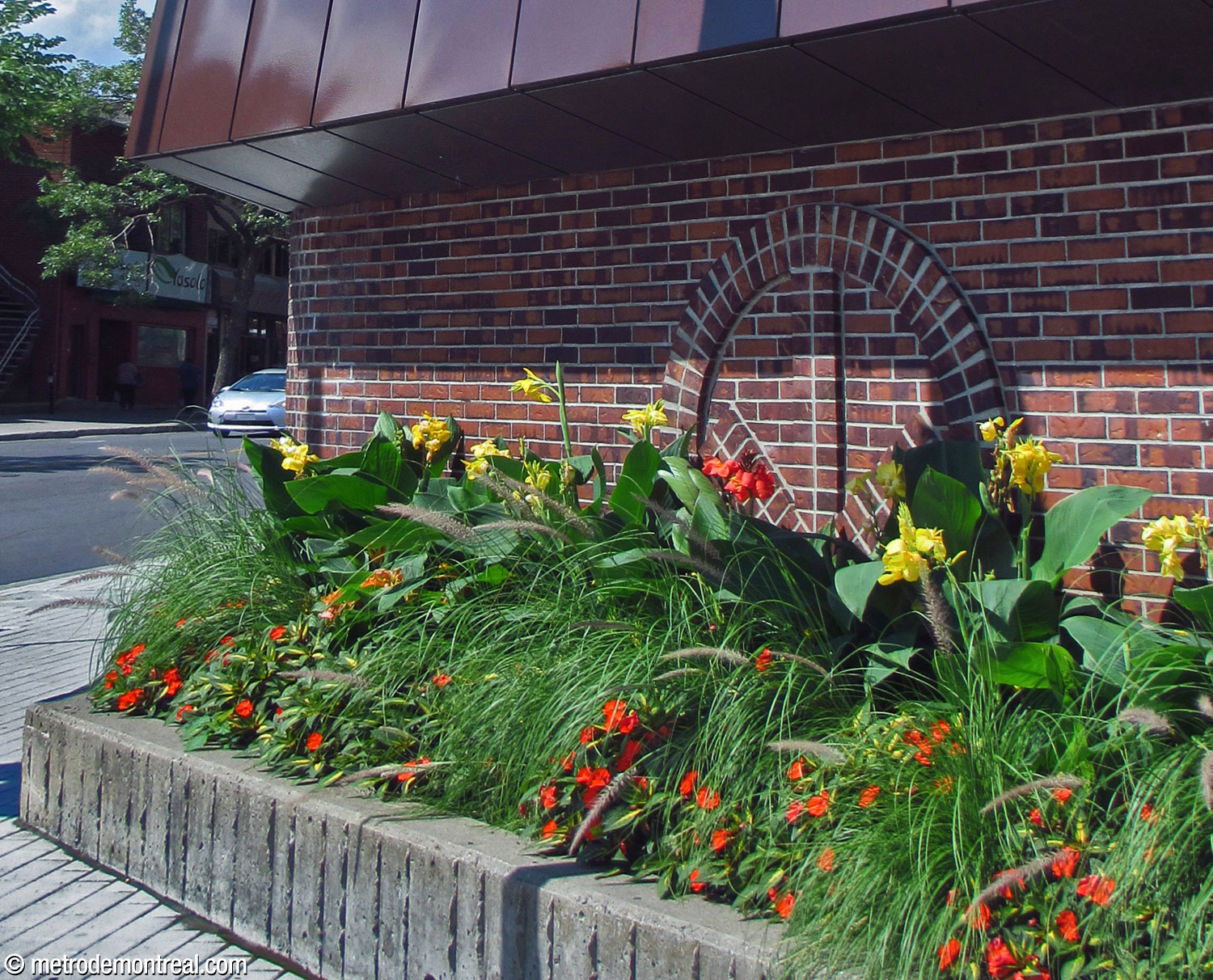 Metro logo design in brick on an exterior wall of Monk Nord headhouse, behind a flowerbed and facing the intersection.