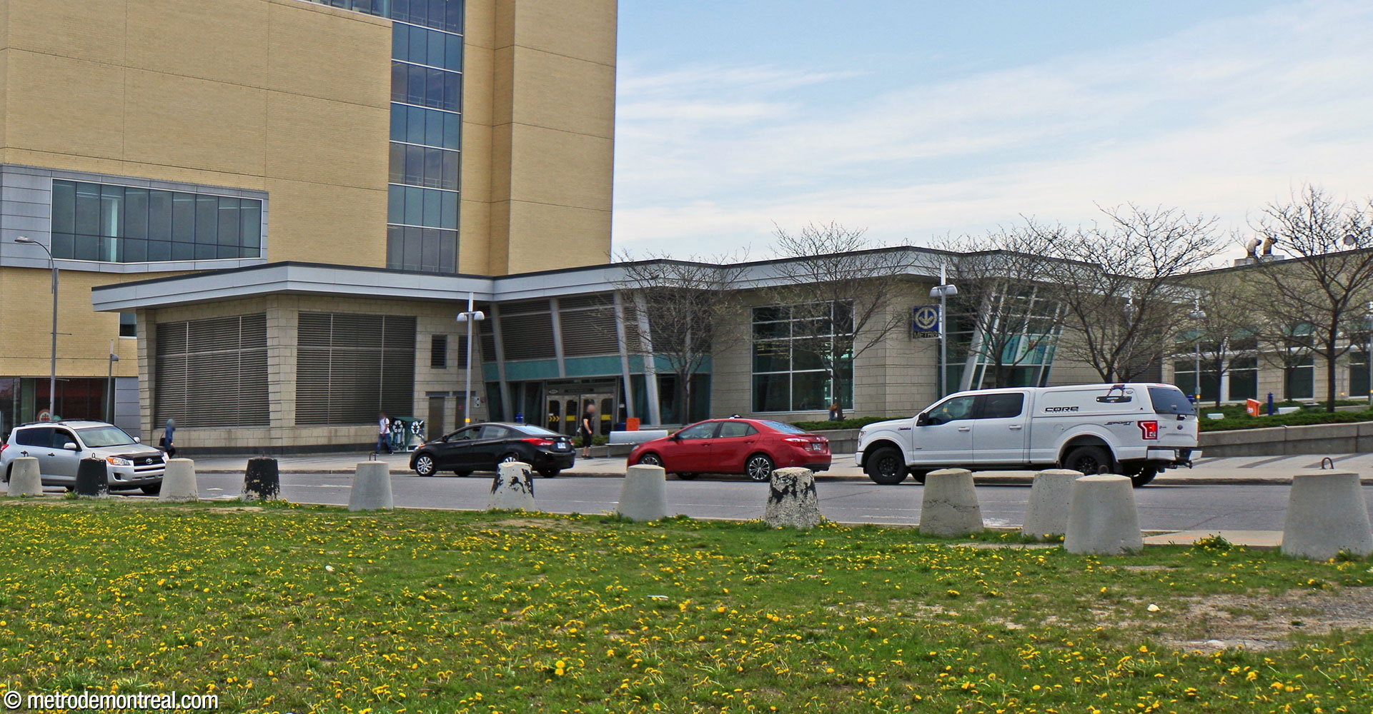 Overview of the metro headhouse. The broad building is clad in pale yellow blocks and features broad windows. The entrances are surrounded by glass and metal porticos leaning outward at the top.