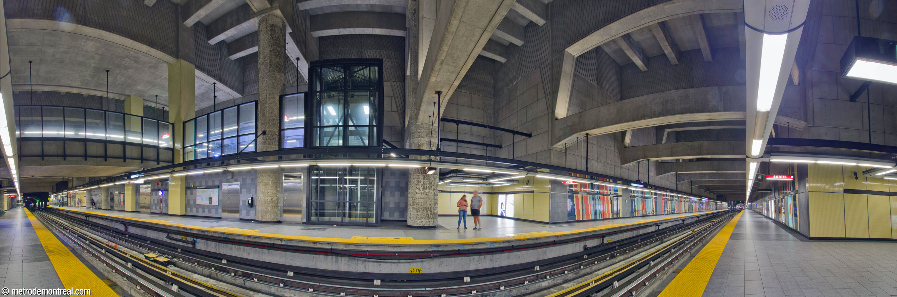 Panorama of the station's vast trainroom, seen from the arrival platform. The trainroom is built in an extremely deep open cut; overhead, a system of huge concrete cross-braces supports the ceiling and walls. At floor level, panels of yellow, red, aqua, dark blue, and black tiling line the walls. The transept and crossover are at left.