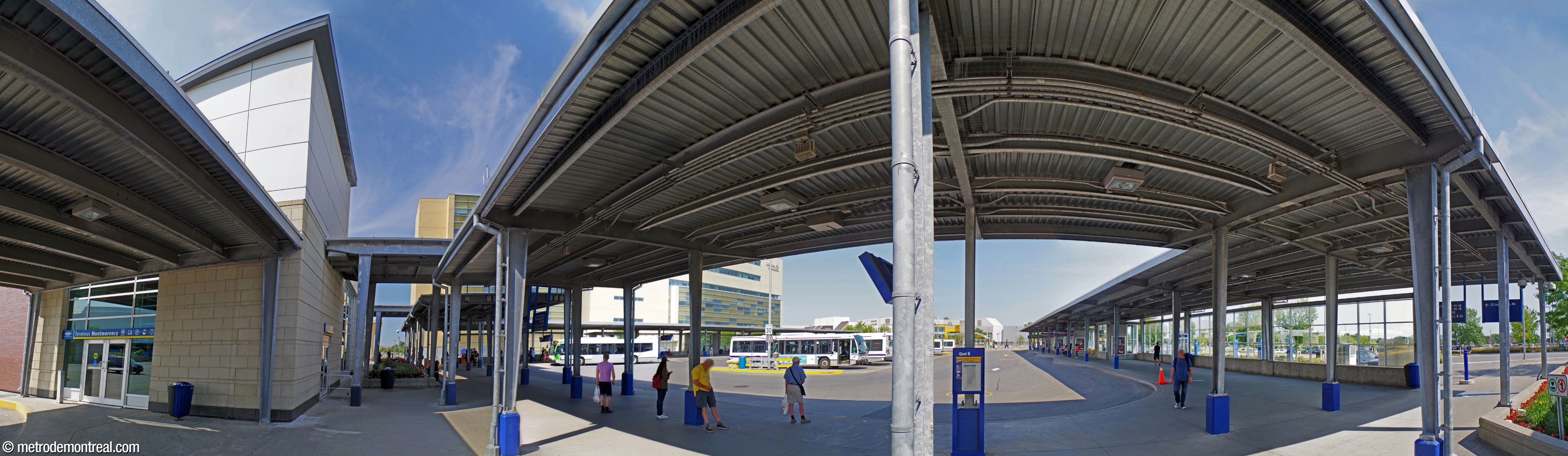 Panorama of the exterior platforms of the bus terminal. The terminal building is at left; its south entrance is visible. The platform areas, including the two jetties framing the bus loop, are open but roofed, with a glass wall on one side.