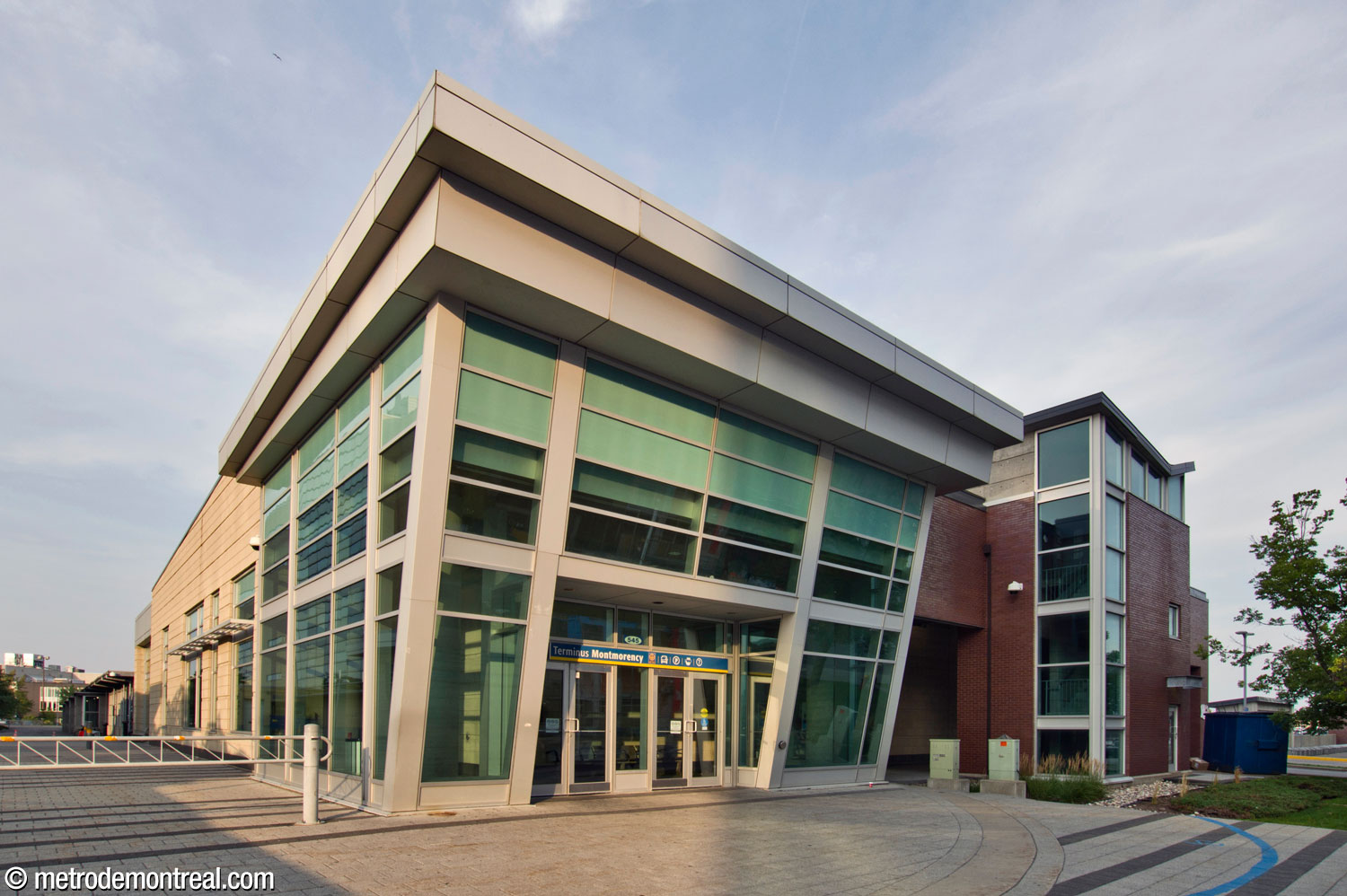The main portico leading into the bus terminal building from the street, consistent with the entrances to the metro station headhouse a few steps away. To its right, a stairwell in the service area is topped by a butterfly roof and repeats the same window layout and glazed red brick material as other stairwells along the perimeter of the adjacent parking garage.