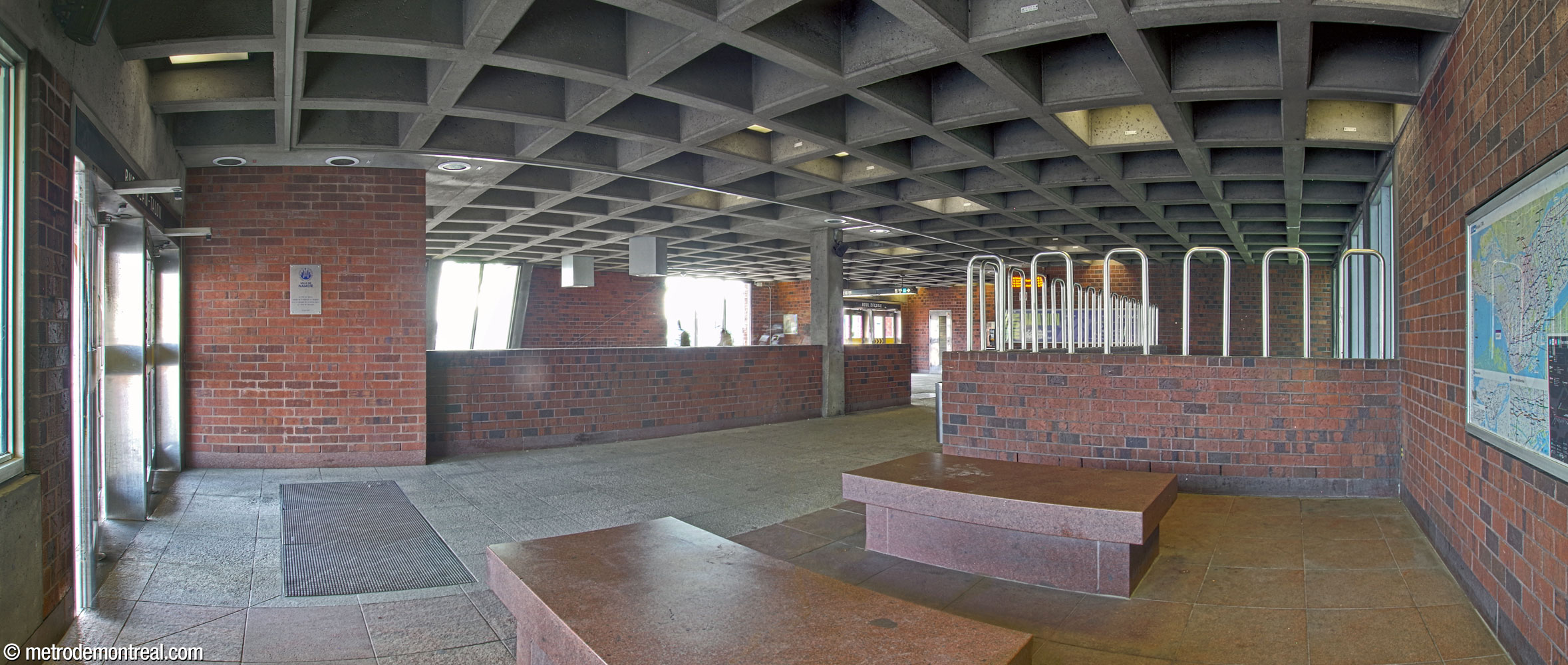 Panorama of the interior of the headhouse, seen from the front, near the Jean-Talon exit. Two stone benches stand in front of the open light shaft. The head of the stairs down to the station is located at the rear, near the exit onto Boulevard Décarie. The wall near the front doors features a commemorative plaque given by the city of Namur, Belgium.