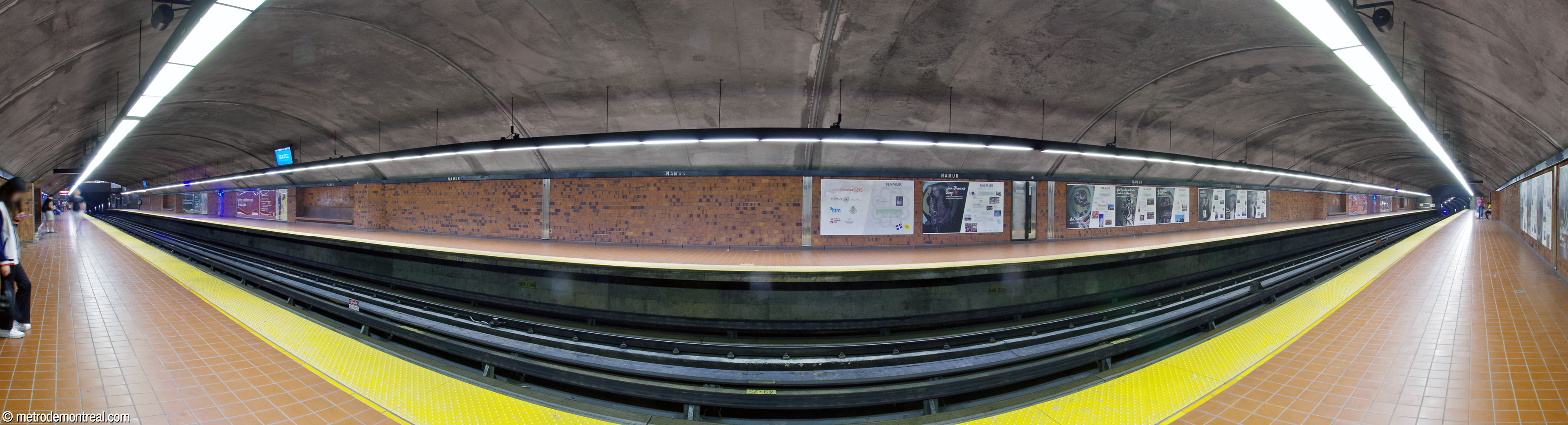 Panorama of the trainroom from the Montmorency platform. The ticket hall is at left, at the northern end of the platforms. The walls are clad in brown brick and the floors in brown ceramic. Informational displays about the city of Namur, Belgium, and its relationships with Quebec and elsewhere line the walls in the middle of the platforms.