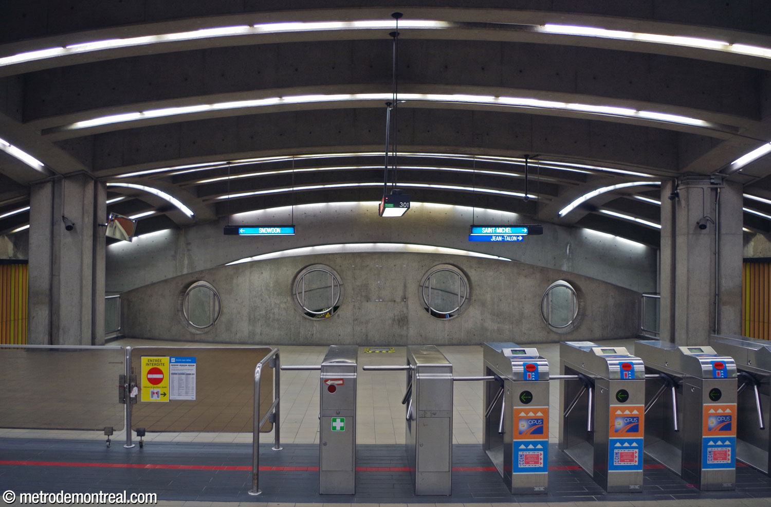 Vue de la ligne de perception dans la billetterie. Derrière elle, le parapet en béton de forme cintrée du côté est de la mezzanine donne sur les quais à travers des hublots.