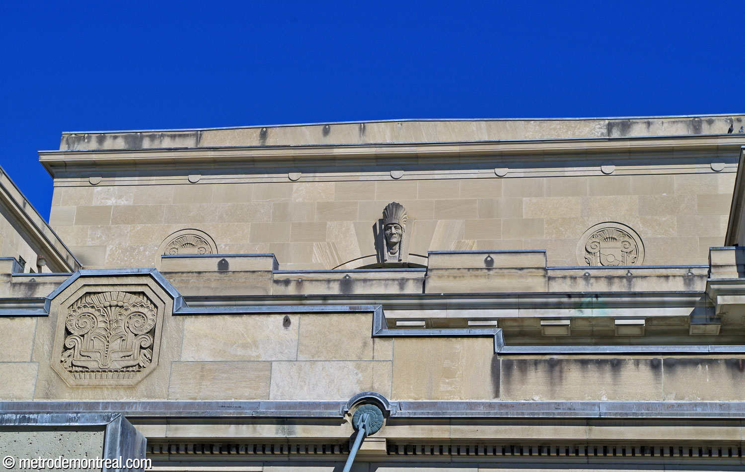 Détail des reliefs décoratifs sur la façade ouest de la gare Jean-Talon, au-dessus de l'entrée du métro. Les surfaces planes sont ponctuées par des médaillons : au niveau supérieur, le buste d'un homme autochtone portant une coiffe fait office de clé de voûte de l'arche d'une fenêtre.