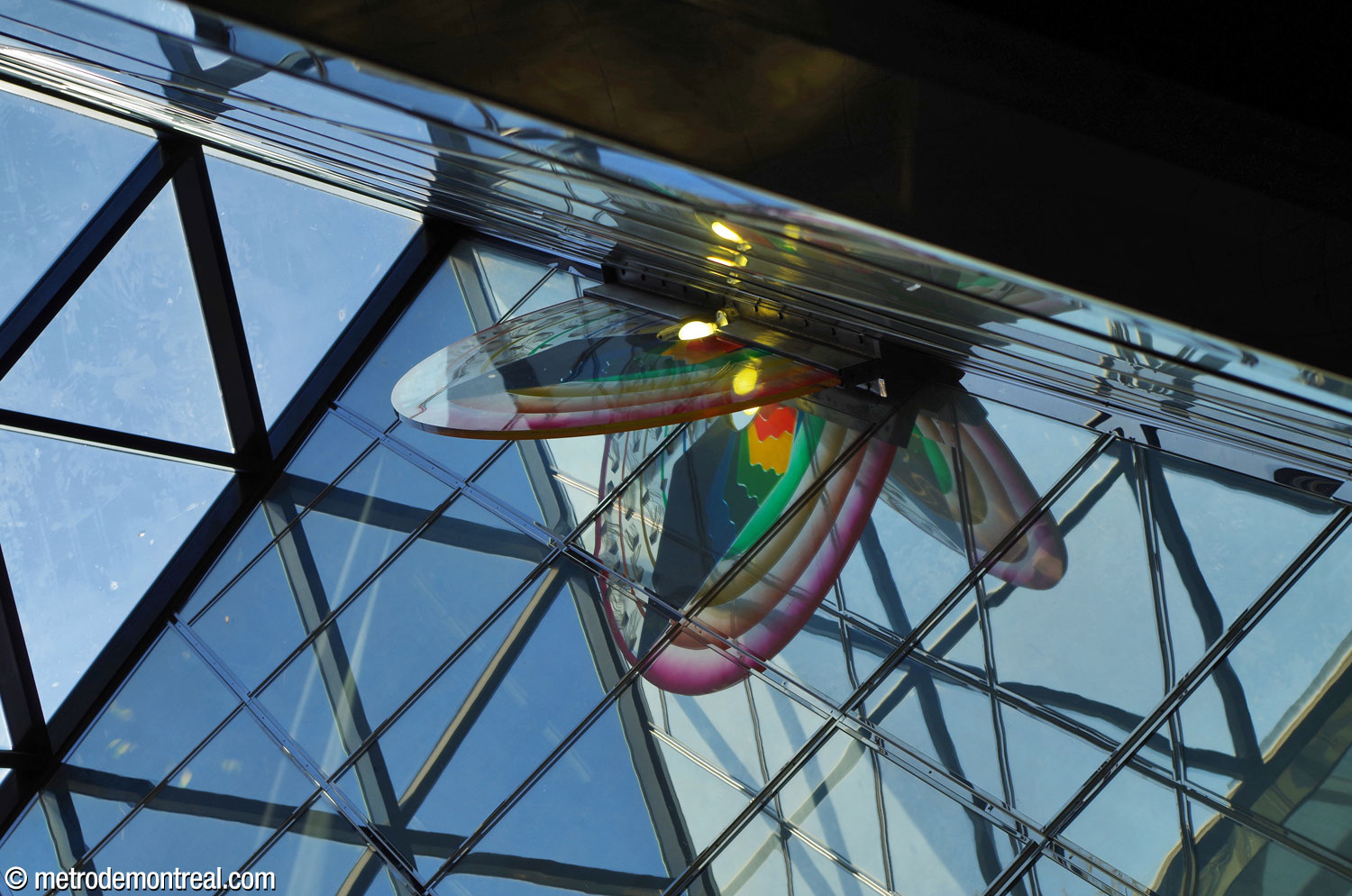 La sculpture acrylique Métamorphose d'Icare, créée par Claire Sarrasin, est reflétée dans les miroirs qui parent les murs à l'intérieur du puits de lumière. L'œuvre de forme semi-circulaire incorpore de vives couleurs arc-en-ciel.