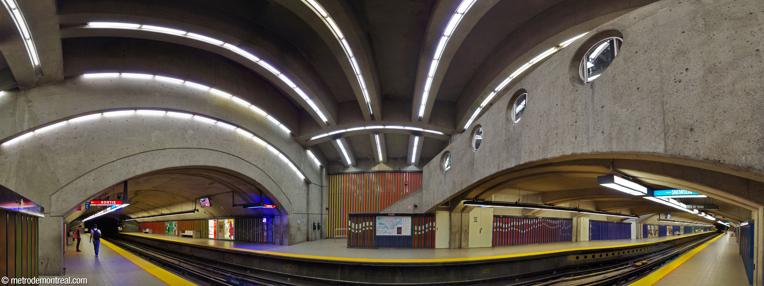 Le grand volume de la station de métro Parc. De part et d'autre se trouvent les portions de la nef construites en tunnel. En haut, le plafond est soutenu par des arches longitudinales et transversales, rehaussées par des bandes de tubes fluorescents. À droite, la mezzanine qui accueille la salle de contrôle est encadrée par un parapet muni de hublots. Les murs des quais et du grand volume sont revêtus de la frise polychrome de Huguette Desjardins, ponctuée par des portes et des bancs blancs.