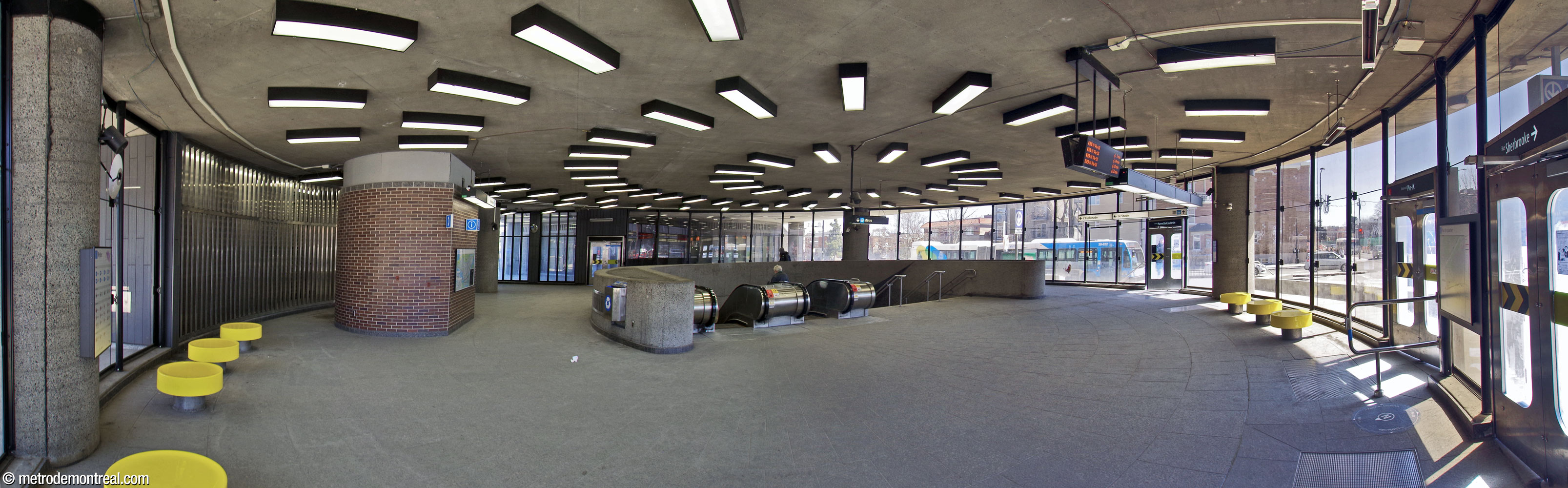 Interior of the rotunda-shaped Pie-IX Est access, with exits to the east (Boulevard Pie-IX, far right), south (Avenue Pierre-De Coubertin, right), and west (Olympic Stadium esplanade, centre left). The staircase to the concourse emerges from an off-centre opening within the room and is surrounded by a round balustrade. Light fixtures on the ceiling emphasize the circular shape of the room and highlight the stairwell. Circular yellow benches line the glass façade.