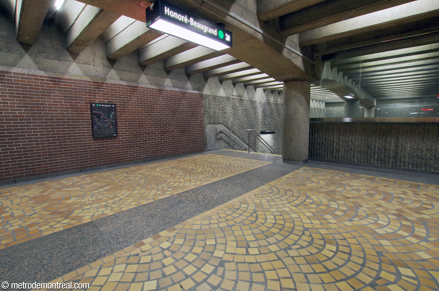 The head of one of the staircases to the mezzanine, showing the station's different finishes: brown brick, fan-shaped patterns of tan floor tiles, and ridged concrete roughened by hammering the ridges. Notice also the pillar-and-arch structure and the X-shaped shadows cast by the arrangement of the light fixtures.