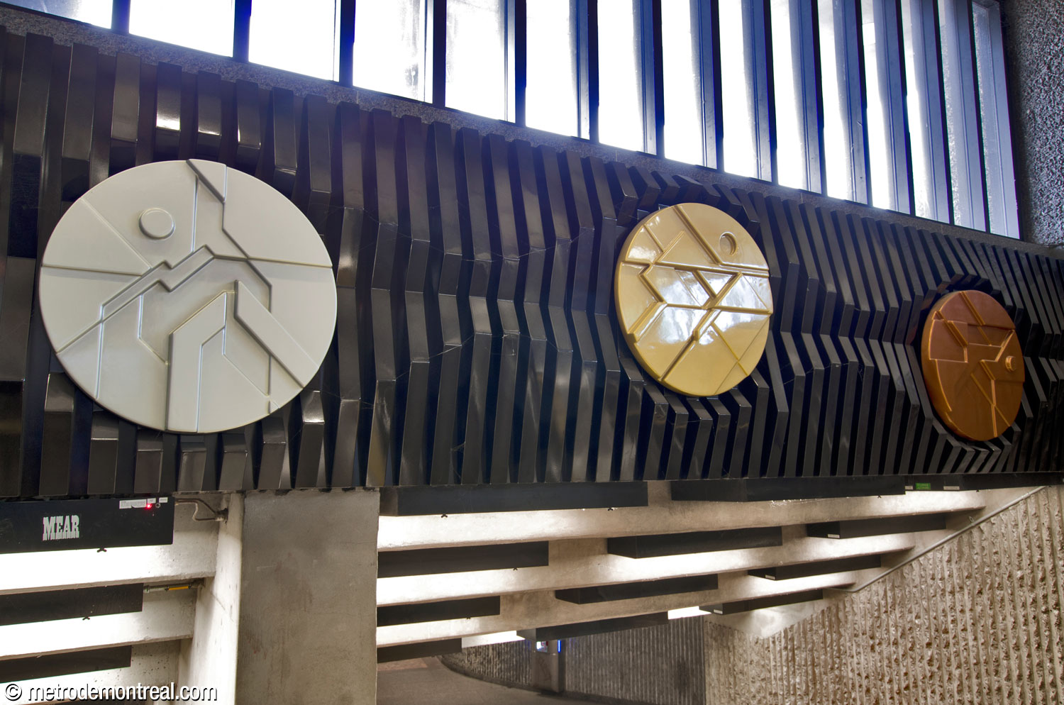 A larger view of the portion of Francis Montillaud’s artwork Point de mire in the west headhouse, featuring three disks coloured gold, silver, and bronze, with haut relief designs suggesting the pictograms that represented the various sports at the 1976 Olympics.