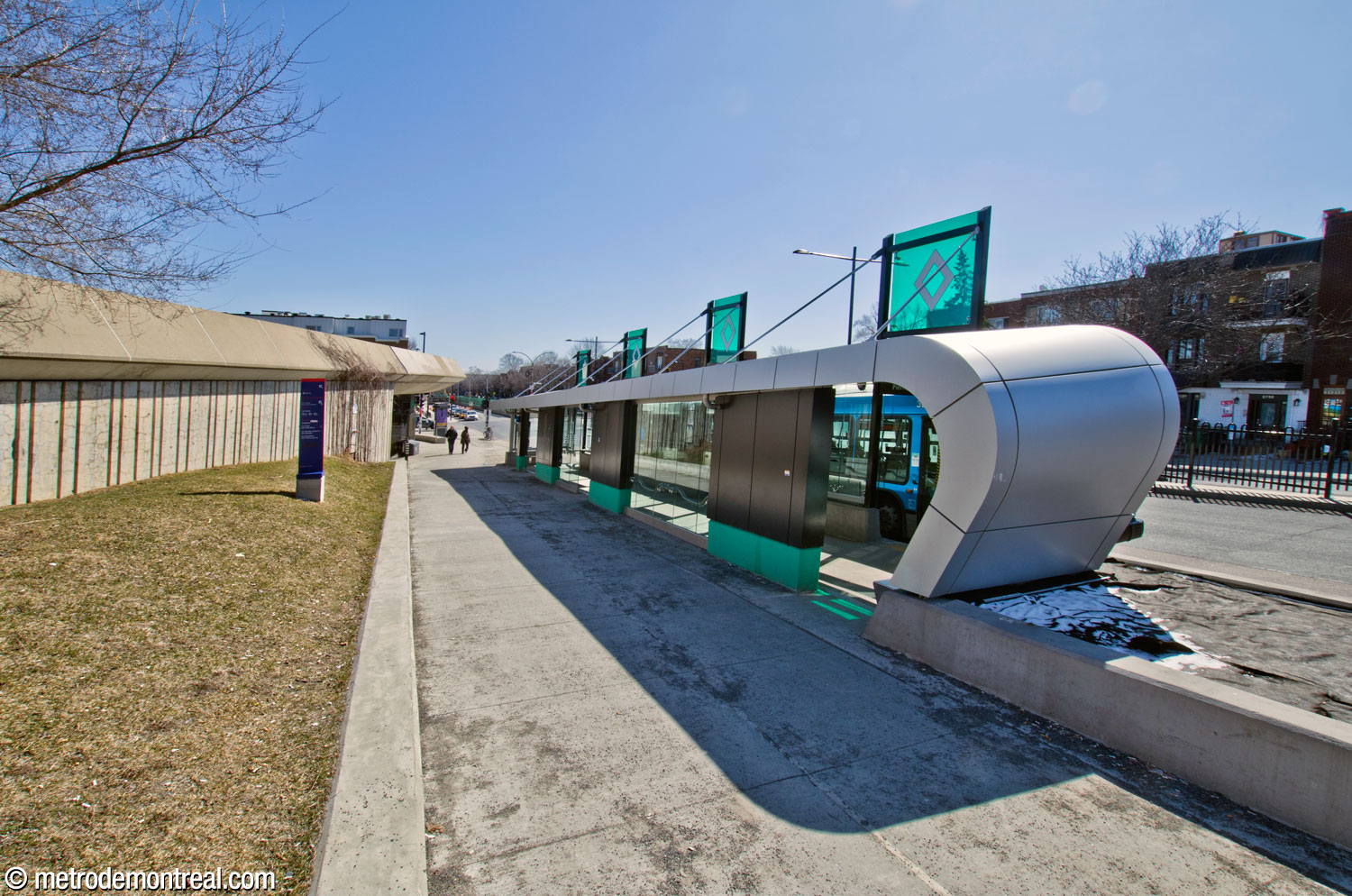 The Pierre-De Coubertin station of the SRB Pie-IX, located immediately north of the Pie-IX Est access. The shelter is inclined with the slope of the street. The metal roof curves down to reach the ground at its north end and is visually supported by sections of wall along the rear. The rest of the walls are glass. Panels atop the roof display the reserved bus lane symbol.