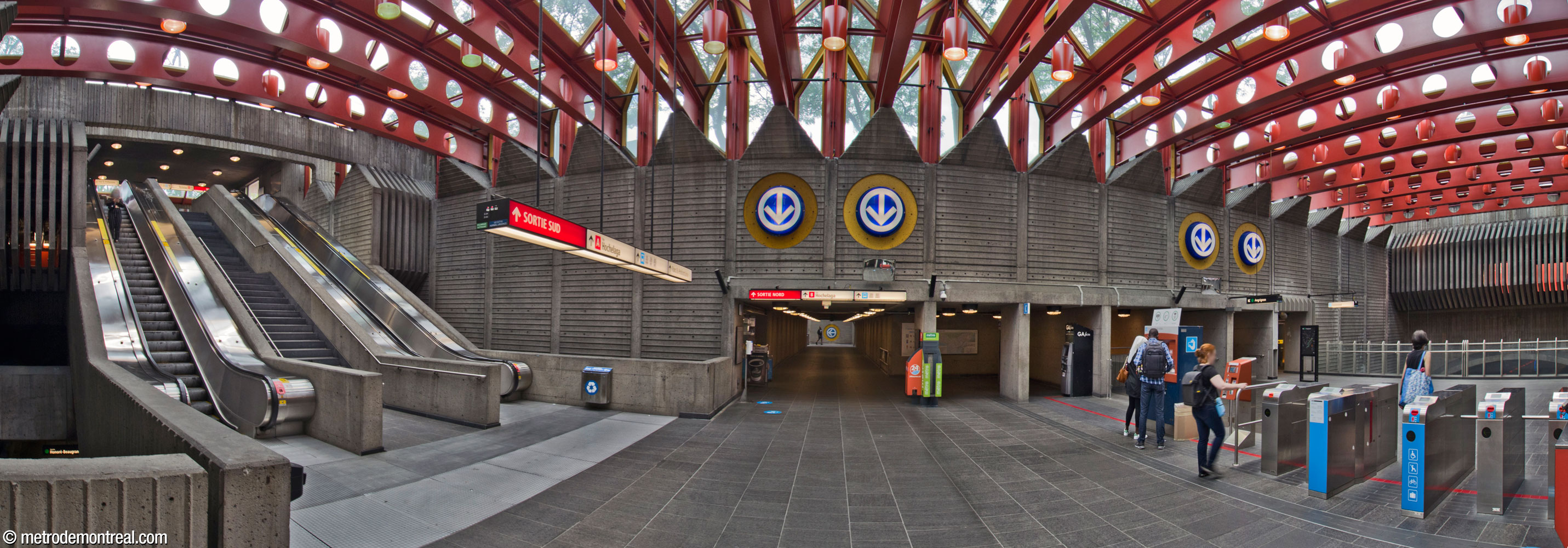 Le mur nord de la billetterie. Ce haut mur de béton est divisé en travées qui correspondent à la largeur des corbeaux qui soutiennent les poutres du toit. Au centre, le tunnel vers l'accès Hochelaga Nord occupe une double travée. À droite, sous les logos décoratifs du métro, les escaliers vers le quai Angrignon sont également aménagés dans des travées.