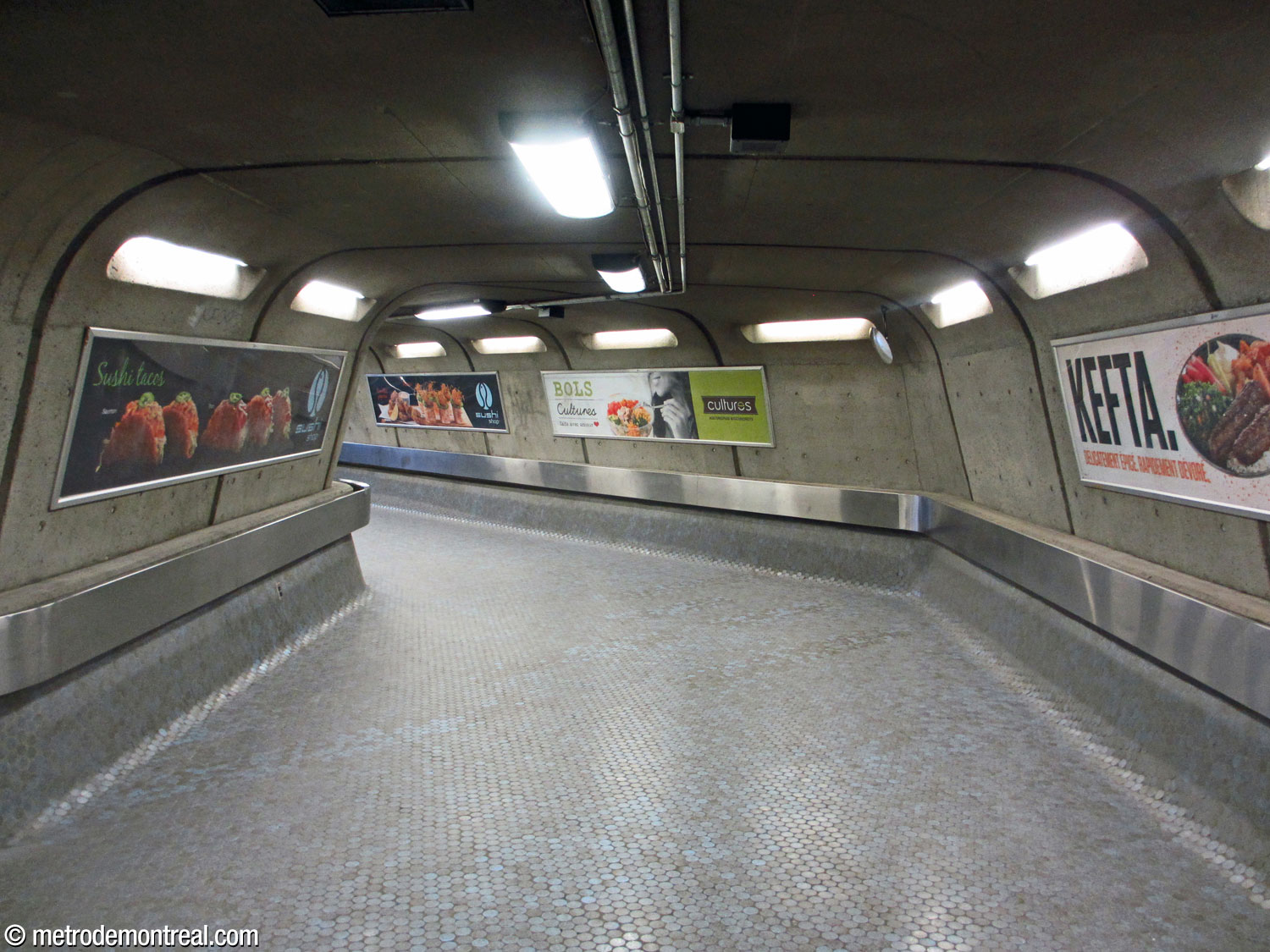 Une section de corridor au niveau de la billetterie. Les murs en béton inclinés vers le centre, le plancher revêtu de céramique, les arêtes courbes, les mains courantes en acier inoxydable et les lampadaires encastrés reproduisent le décor du grand volume.