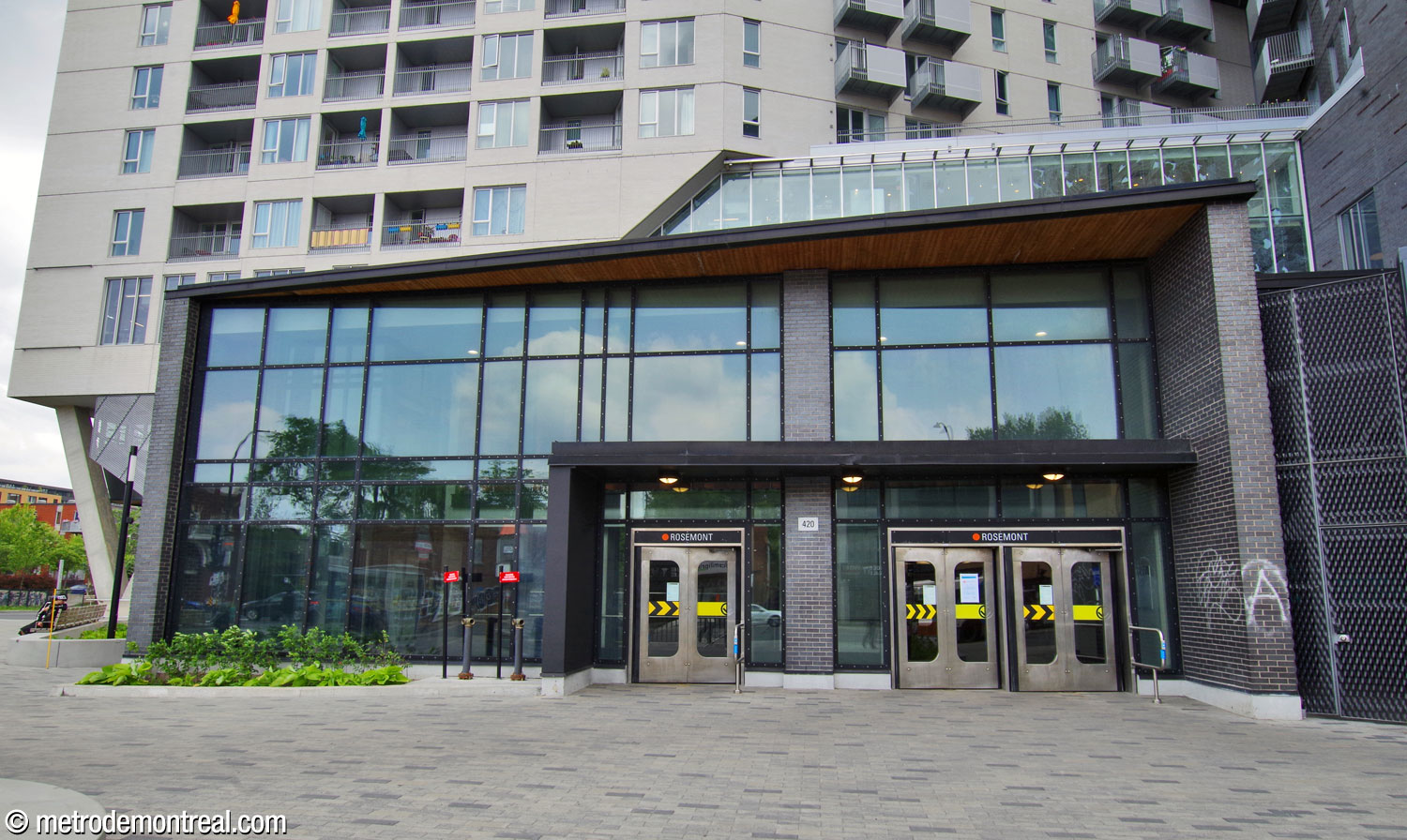 La façade principale de l'édicule, face au boulevard Rosemont, entouré par la tour de logement social. La façade que l'on voit ici est constituée principalement d'un agrandissement de deux étages, construit en 2017 pour recevoir l'ascenseur vers la billetterie. La brique foncée et le verre teinté sont en continuité avec l'édicule existant, mais la saillie oblique du toit est accentuée avec un revêtement en bois.