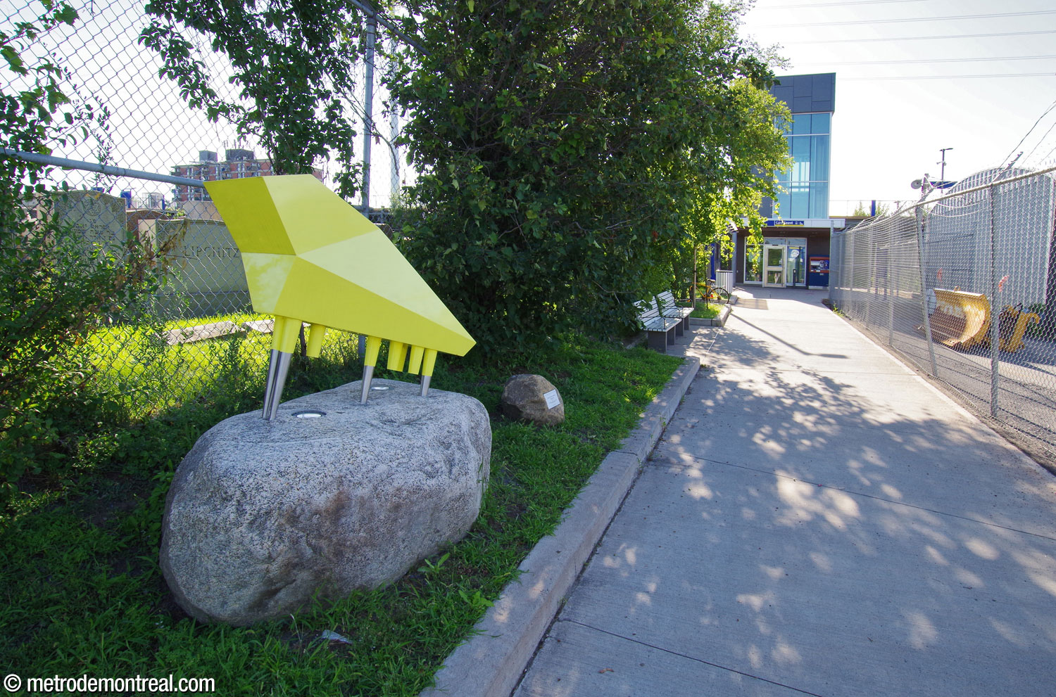 La sculpture Déplacement de Marie-France Brière est située à côté du passage piéton vers l'édicule de la gare (au fond). La forme abstraite en métal jaune représente une chaussure athlétique, en symbole de mobilité.