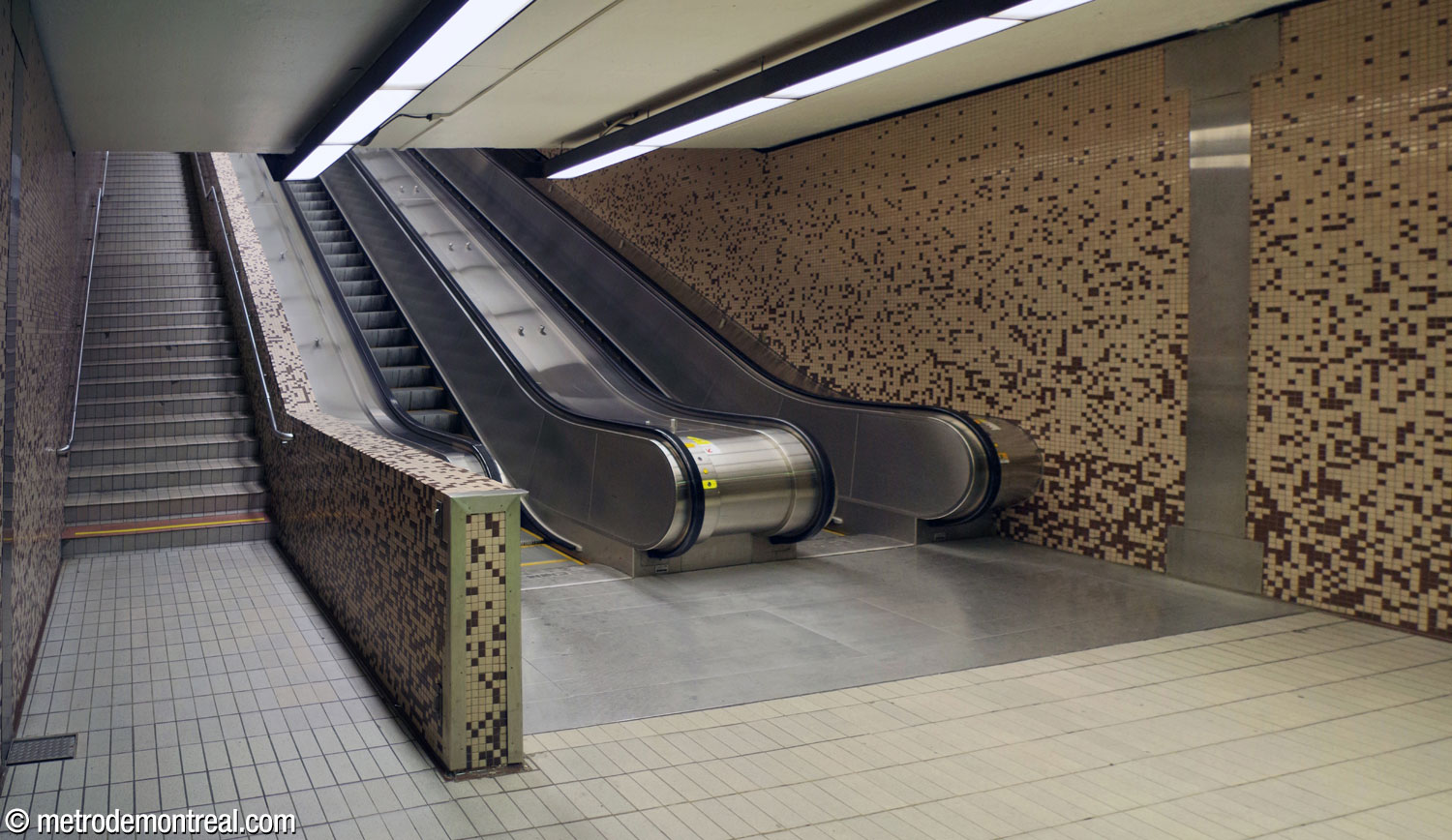 Le pied de l'escalier montant du transept vers la billetterie. Élément distinctif, les escaliers mécaniques sont séparés de l'escalier fixe par un muret revêtu de la même mosaïque beige et brun que les murs.