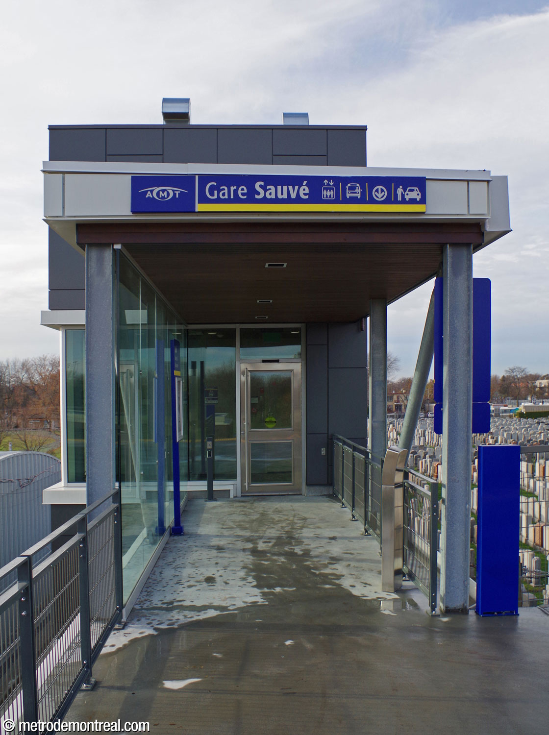 Une passerelle à construction partiellement ouverte (recouverte par un toit et vitrée d'un côté) relie le quai à l'étage de l'édicule de la gare.