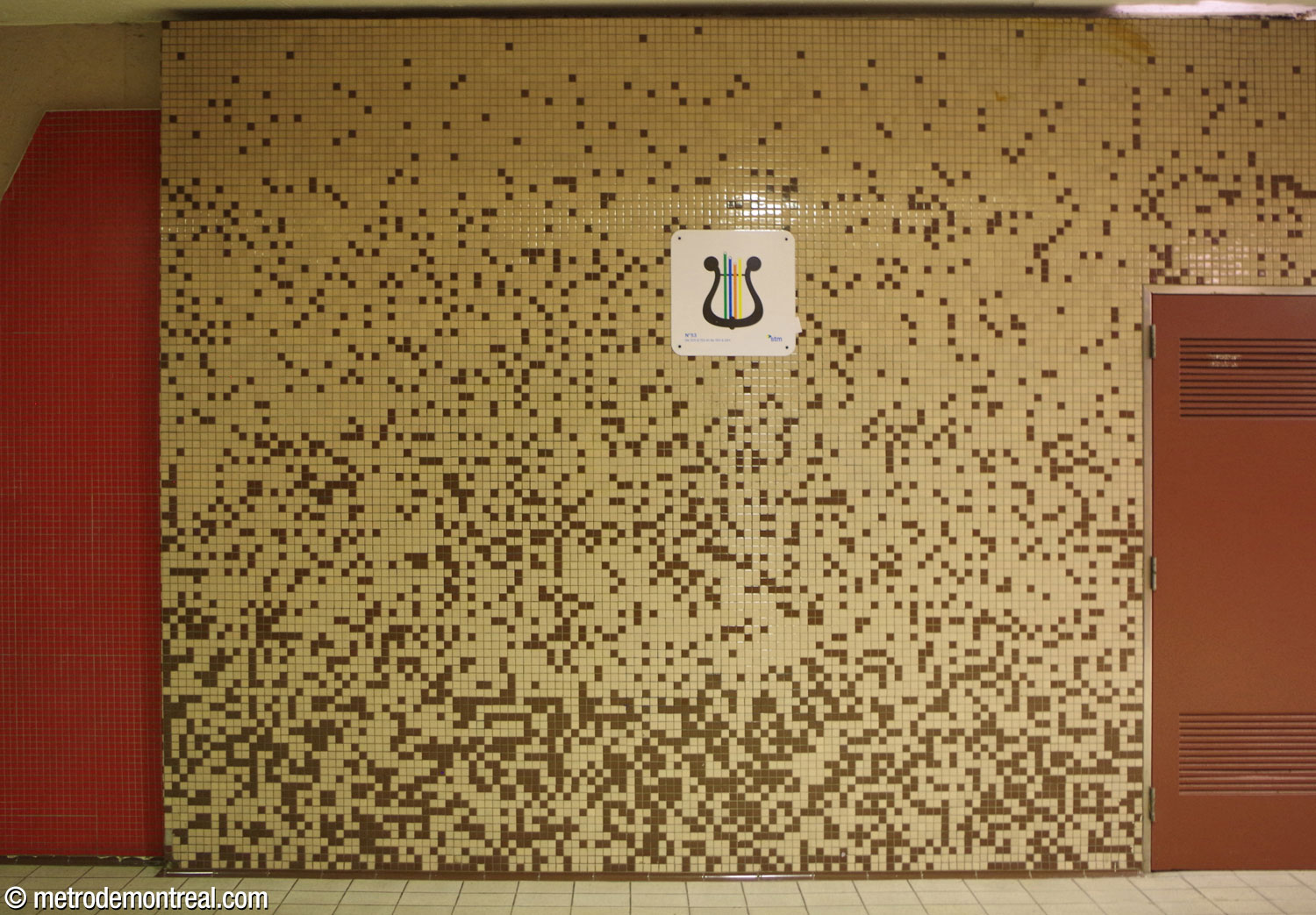 Une section de mur au niveau du transept présente le dégradé de brun sur fond beige partagé par les finitions murales mosaïques dans toutes les parties supérieures de la station. À gauche, une partie de l'un des murs carrelés de mosaïque rouge qui encadrent les percées visuelles entre la passerelle et les quais.