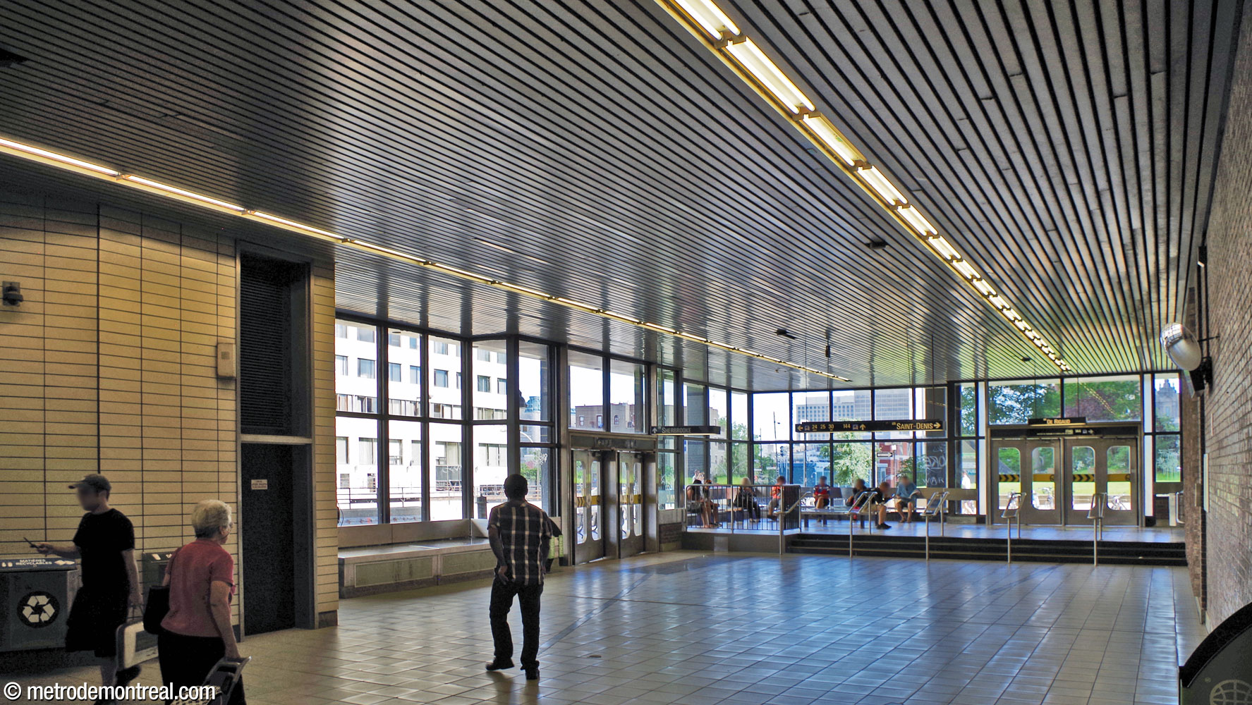 View toward the south of the Berri Ouest entrance, with large expanses of glazed curtain walls looking out onto the bus stops on Rue Berri and Rue de Rigaud. A waiting area for bus passengers occupies the corner.