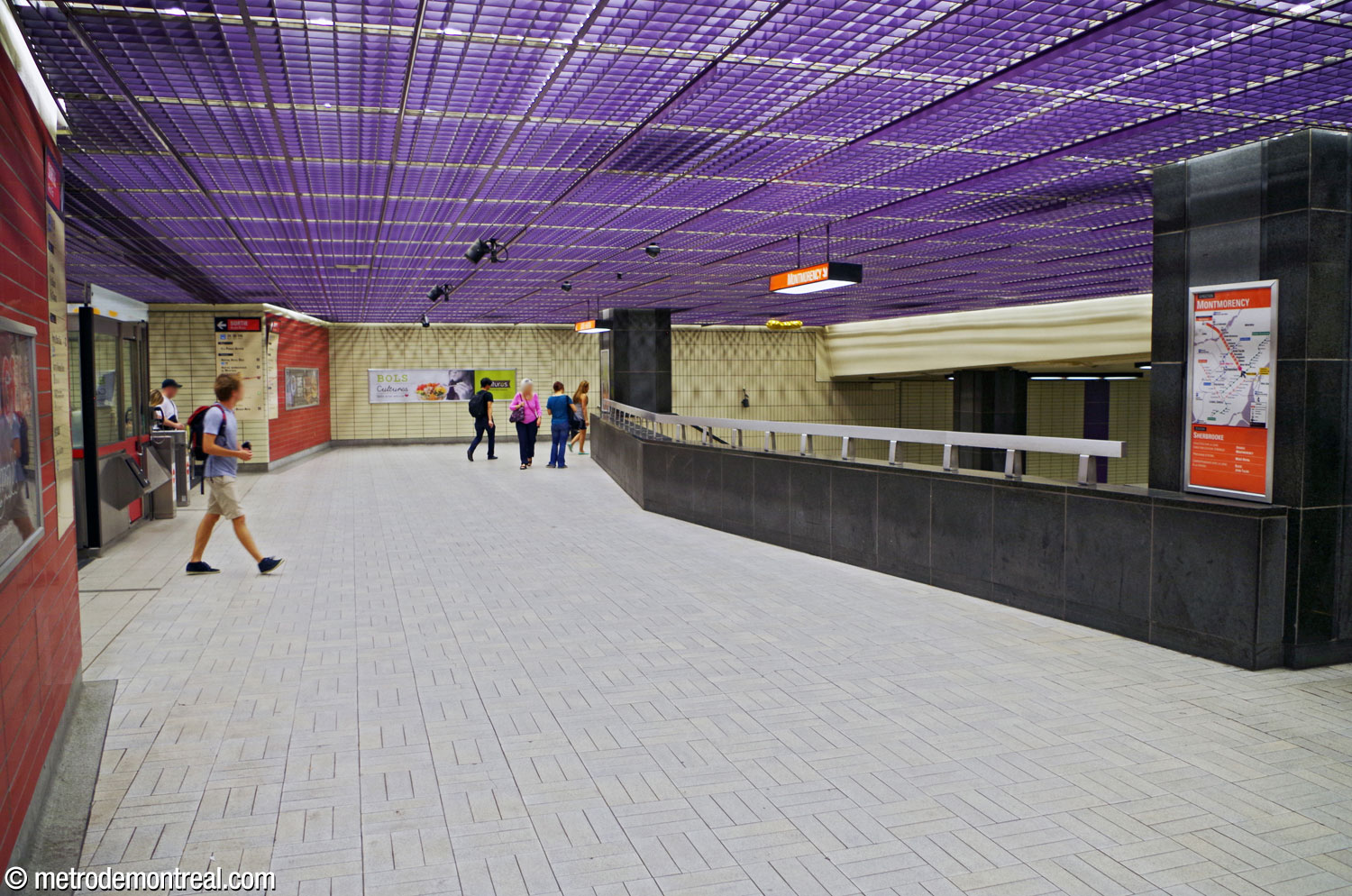 The fare-paid zone overlooking the platforms. Overhead is the bright purple drop ceiling grille. Orange accent walls are found at either side of the gateline (left), breaking up the pale yellow tiling. The parapet is in the same black stone as the columns.