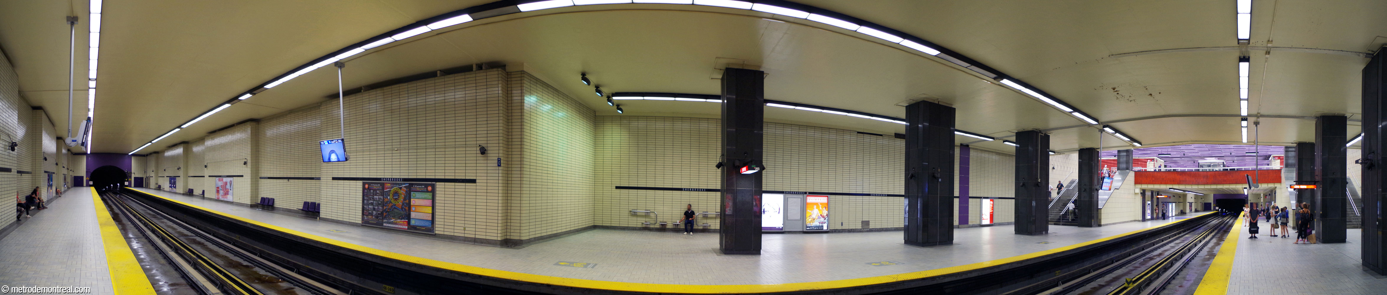 Panorama of the trainroom, built cut-and-cover. Alongside both platforms, long, broad side aisles (centre right), set off from the platform by large black columns, lead to the staircases to the mezzanine (right). The walls are clad in pale yellow tiles throughout.