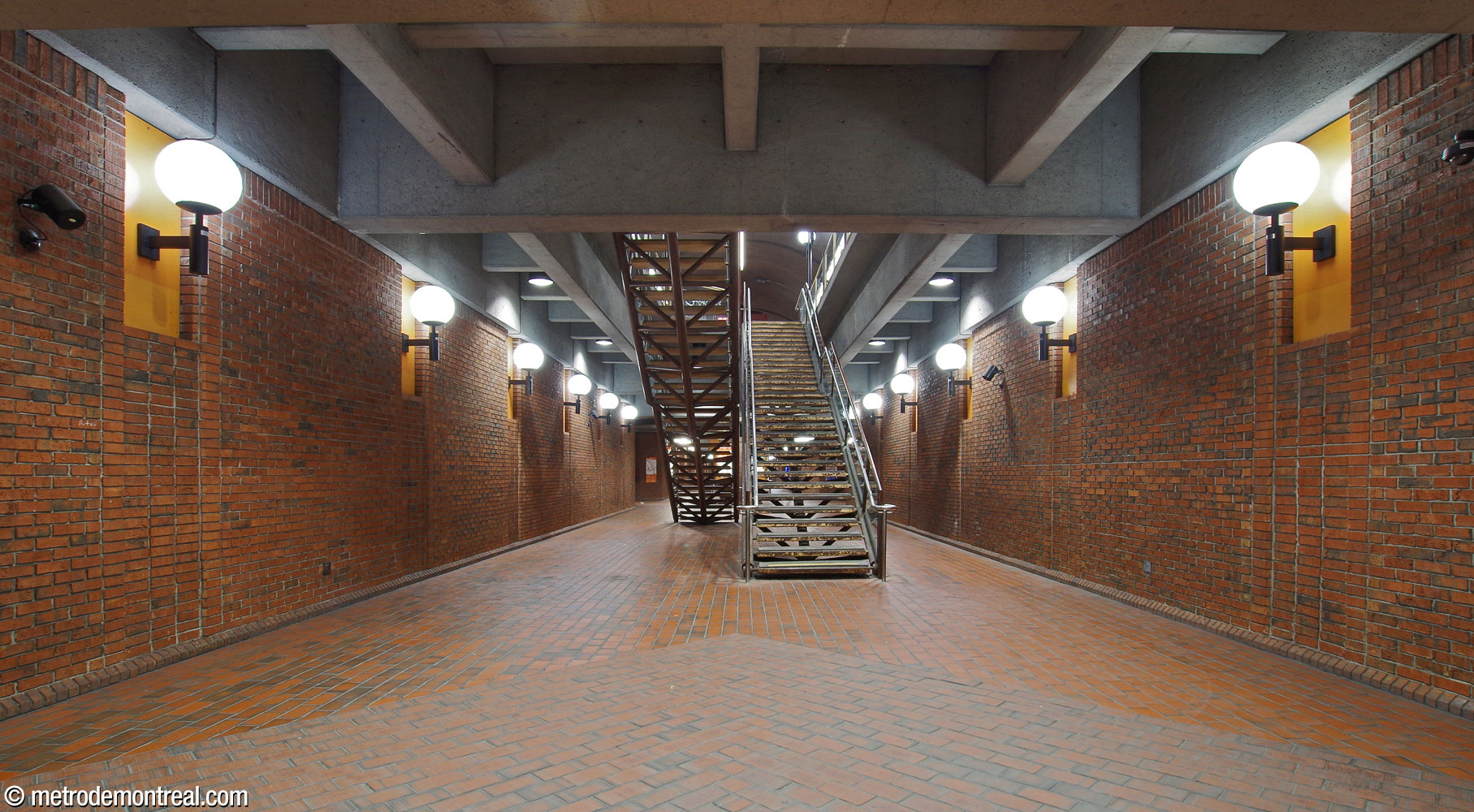 The lower level of the central vault, looking westward from the eastern end. The corridor is lined with a row of wall-mounted spherical light fixtures mounted in recesses with yellow accents. Note also the various arrangements of brickwork and floor tiles.