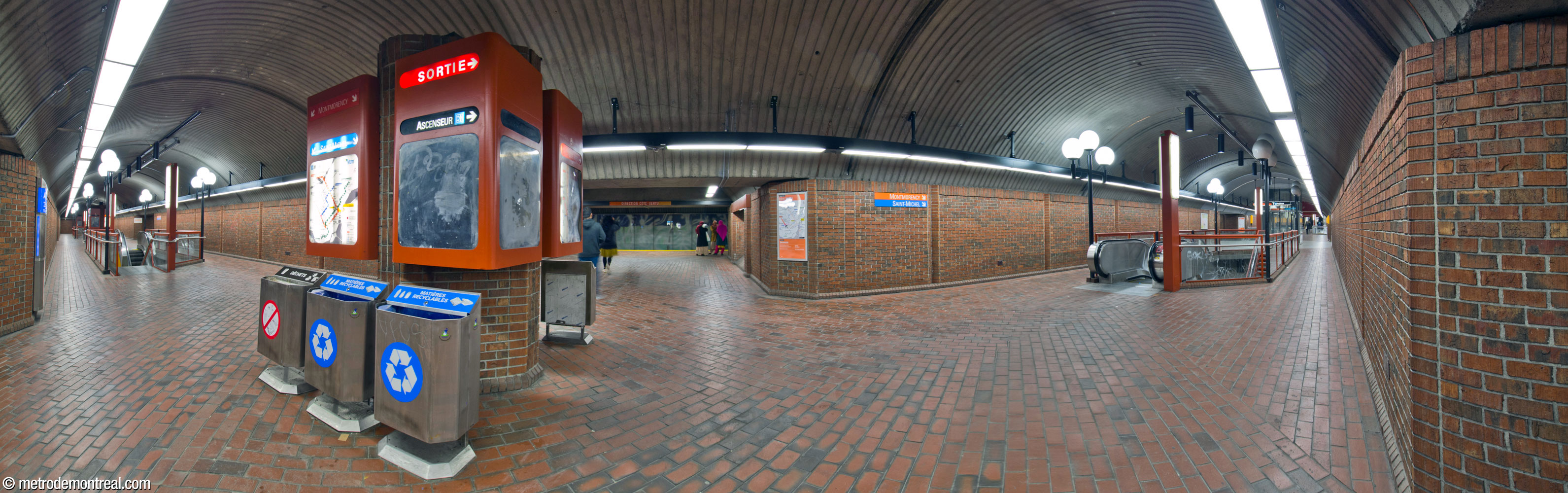 Lengthwise panorama of the upper level of the central vault of Snowdon metro station. The exit is at the end of the vault at far right; at centre, one of the transverse tunnels leads to the Côte-Vertu platform. Along the centre line of the vault, stairwells containing paired staircases and escalators lead to the lower platforms; lamp posts with triple spherical lamps stand at the corners of the stairwells. At the intersection between the central vault and each transverse tunnel stands an information pillar with line maps.