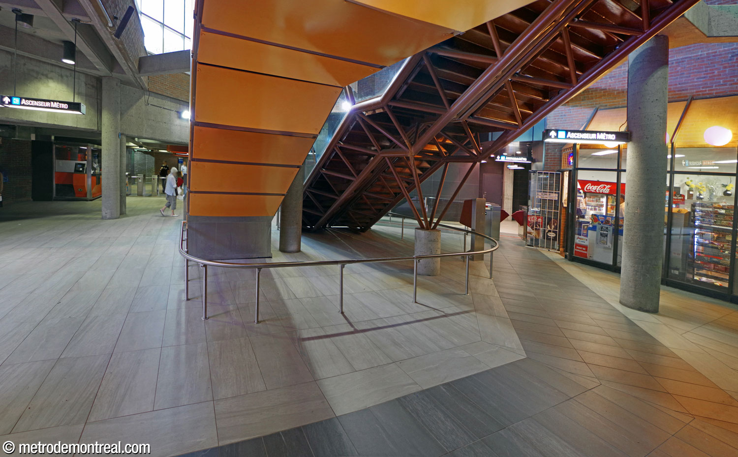 View toward the gateline from underneath the stairs to the exit. Unusually, the design of the ticket hall left this broad space outside of the path of traffic flow. It now offers a path (marked by dark grey tiles) between the elevator to the exit and the ticket gate giving access to the elevator to the platforms.
