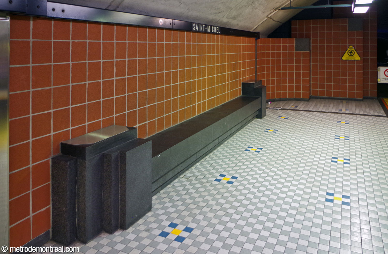 A platform bench in black granite with stepped end pieces. The walls are clad in terra cotta blocks and the floor in white and grey chequered tiles with regularly spaced blue and yellow decorations. At the rear, stepped curved panels of terra cotta and granite blocks mark the platform end walls.