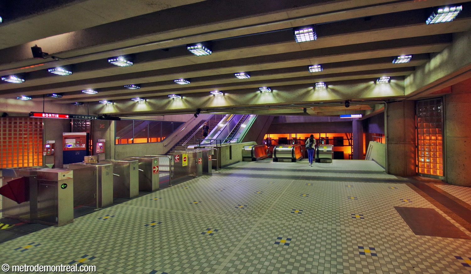 The ticket hall, looking northwest from the fare-paid zone. The stairs to the east headhouse are behind the turnstiles at centre; at centre-right are the stairs to the platforms. The orange glow seen through the glass block walls and windows and in the space at rear is from the large light fixtures in the central volume.