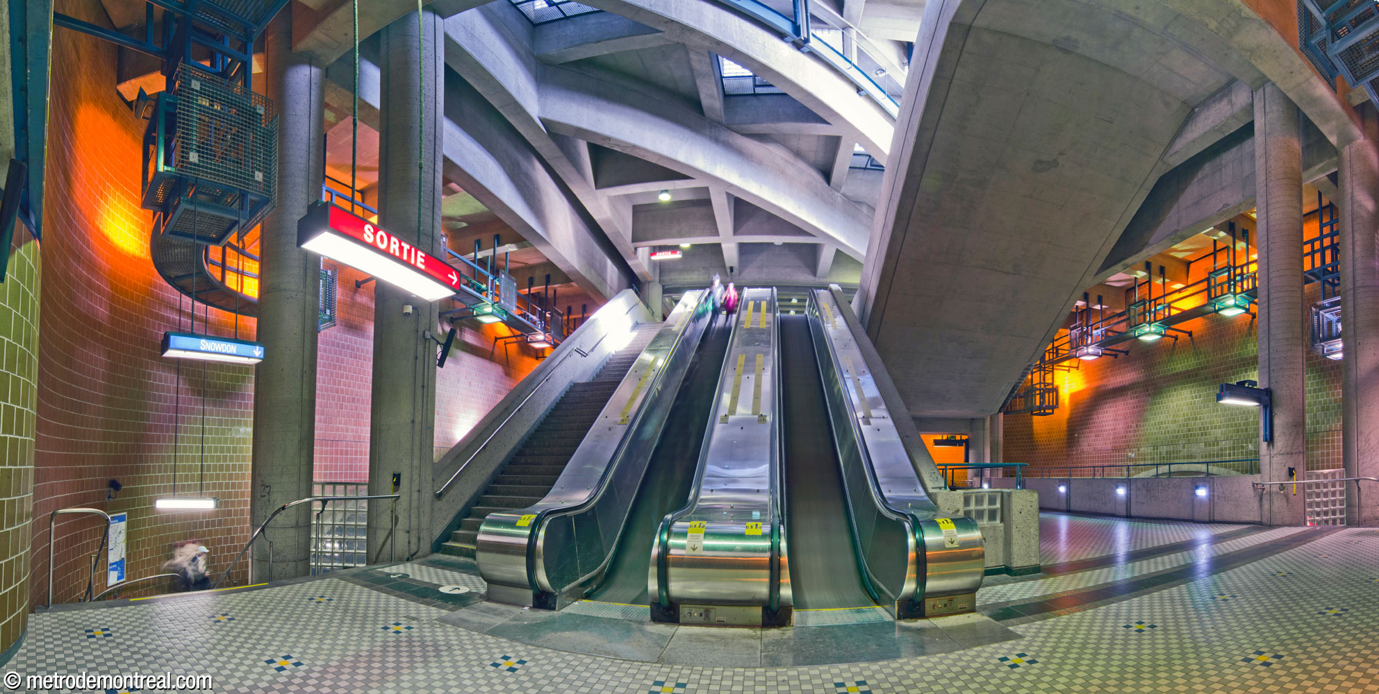 The central volume of Saint-Michel station, seen from transept level at the landing at the foot of the stairwell to the ticket hall. To the left and right, stairs lead down to the departure platform; at right is the crossover to the arrival platform. At upper right is the stairwell to the Saint-Michel Est headhouse, whose windows admit natural light into the volume. The station's unique cube-shaped light fixtures are overhead at left and right. The volume, and the headhouse above, are supported by four pairs of pillars, two pairs of which are visible at foreground left and right.