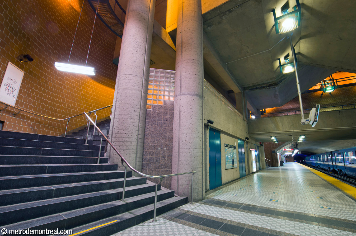 The eastern flight of stairs going up from the arrival platform to the footbridge, at right. The stairs wrap around one of the four pairs of pillars that support the roof of the cube-shaped main volume. An accent of glass and rose granite blocks joins the two pillars.