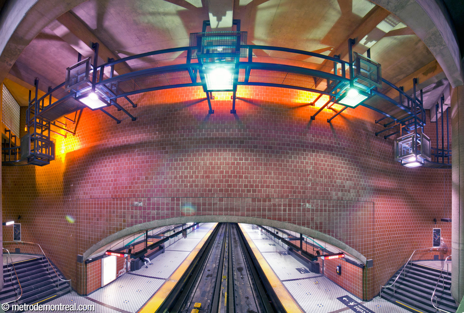 Panoramic view of the high, flat spandrel wall on the west side of the main volume, above the trainroom vault. Above, the station's impressive cube-shaped light fixtures are installed on a railing and catwalk running along the contour of the volume, and cast dramatic shadows onto the ceiling.