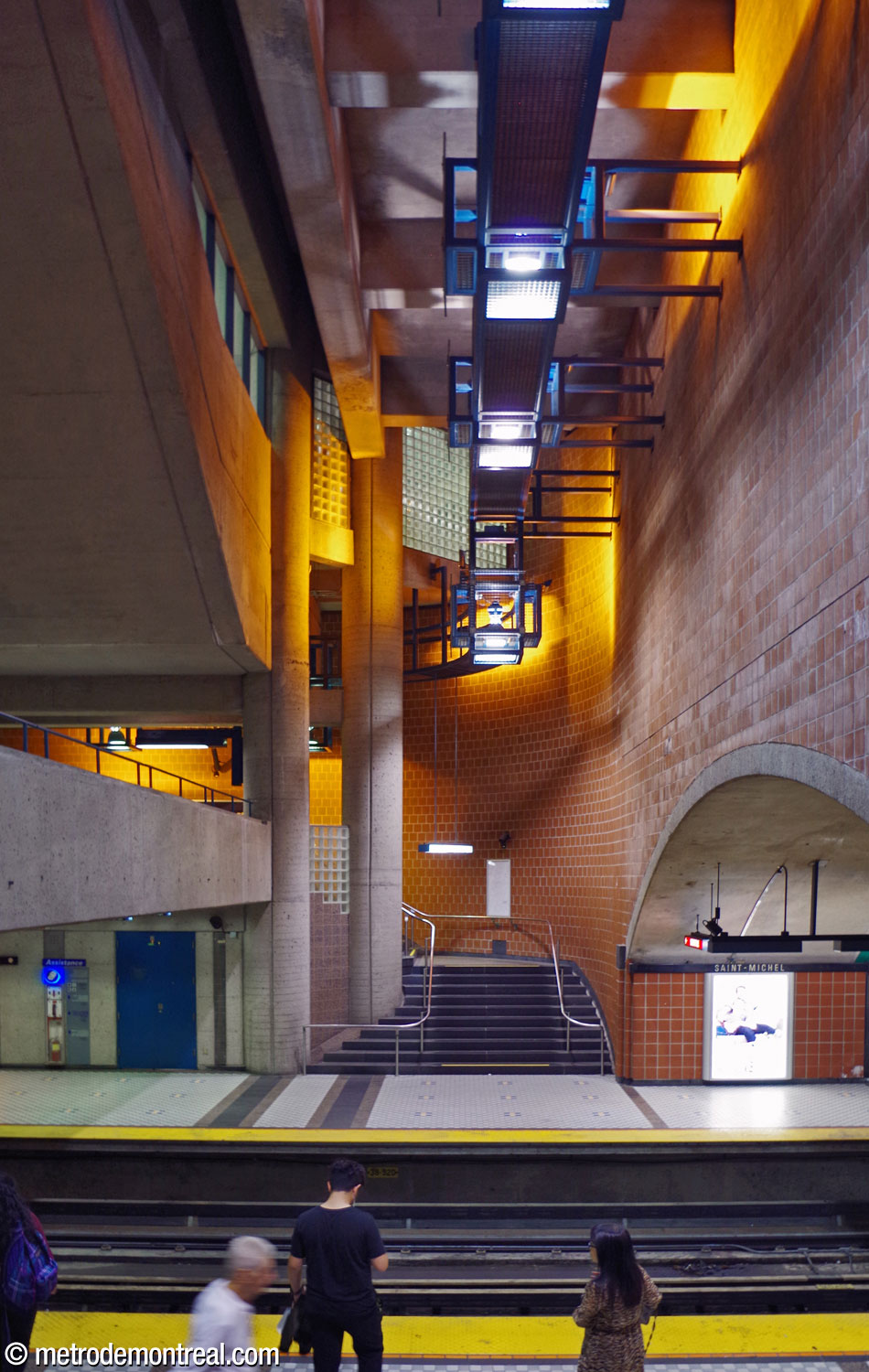 Looking across the tracks from the departure platform stairs. The edge of the crossover (left) and the spandrel wall above the trainroom vault (right) define a high vertical space housing stairs up to the transept level (centre). The station's distinctive light fixtures are installed along a rail passing through the axis of this space. Behind them, windows and glass block walls admit their light into the ticket hall.