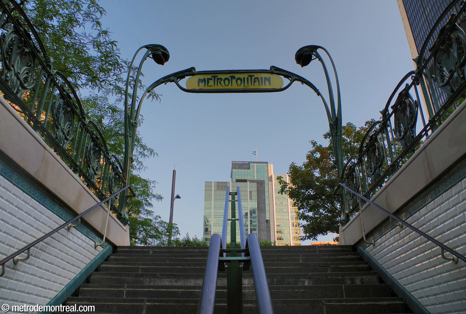 Vue en contre-plongée de l'entourage Guimard, avec l'emblématique enseigne « Métropolitain » entre les élégants luminaires « brin de muguet ». Pour encore plus d'authenticité, l'entrée est revêtue des carreaux biseautés blancs typiques du métro parisien.