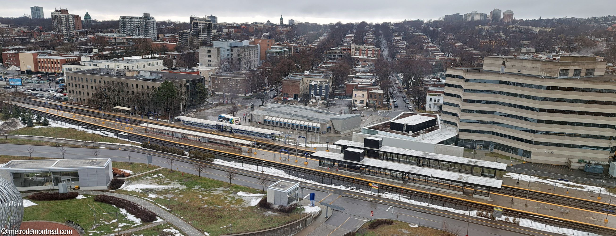Overhead view of the station complex seen from the MUHC. The original headhouse is at centre, surrounded by a bus loop; the new headhouse is to its right, adjoining another building. Closer to the foreground, the commuter train station platforms traverse the image, cutting the station off from the hospital campus; two tracks pass between the platforms, and a third track passes south of the station, turning the southern platform into an island platform. The older train station shelters stand across from the original metro headhouse at left, with the newer International-style shelters across from the new headhouse at right. In the bottom left is the MUHC campus headhouse, at the end of the tunnel from the original headhouse.