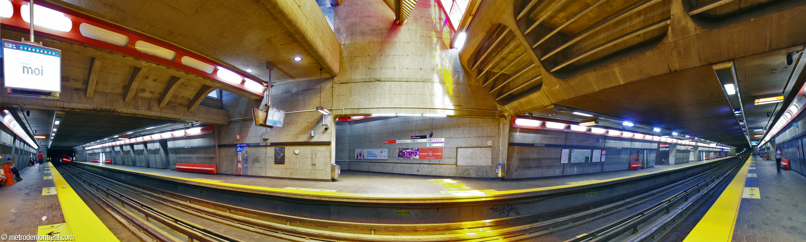 Panorama of the trainroom from the Montmorency platform. The walls are in exposed concrete with red-orange benches and light housings. The station's main volume, within the original headhouse, is at centre, as is the stairwell to that headhouse; the stained glass window and sculpture by Marcelle Ferron are overhead, admitting brightly coloured light into the volume. The accesses to the new headhouse are further down the platform, at right.
