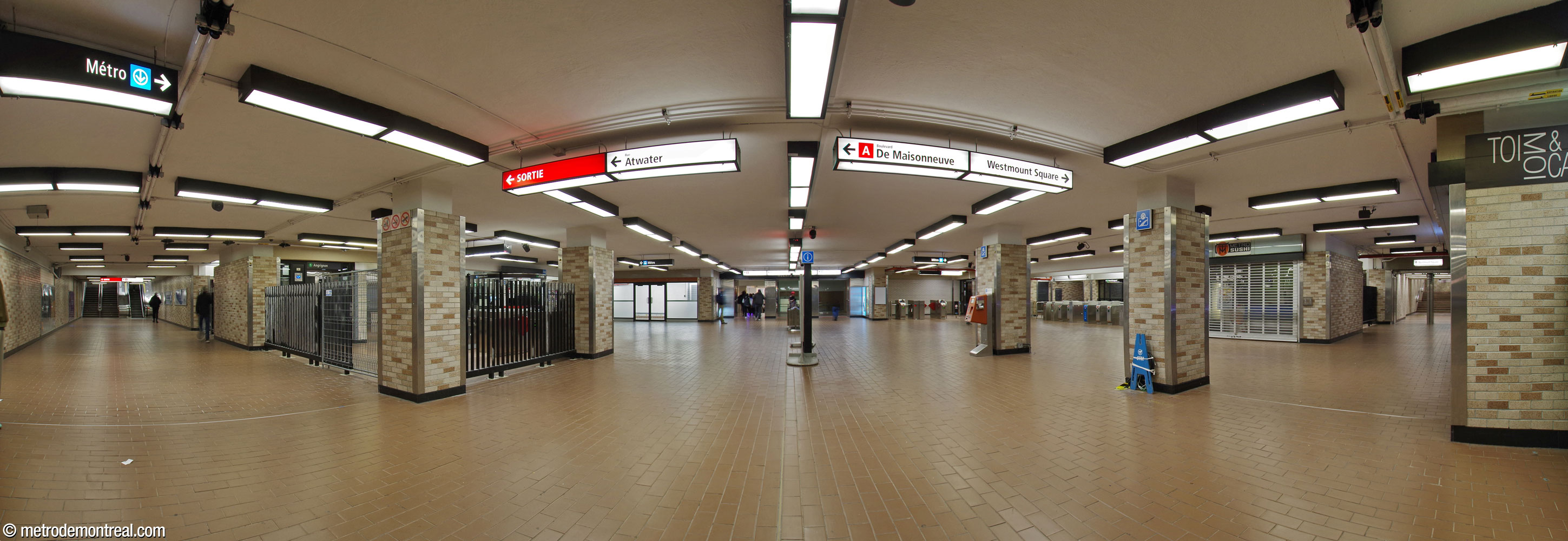 View of the concourse, facing south, toward the entrance to Complexe Alexis Nihon. At left, a corridor leads to the De Maisonneuve exit; at right is the entrance to the long tunnel to Westmount Square. At centre-left, the fare-paid zone for the elevator to the Angrignon platform, enclosed by black grilles, protrudes into the concourse, extending the corridor toward the De Maisonneuve exit. The elevator to the Honoré-Beaugrand platform is in the background at centre-right, in a bay clad in white brick.