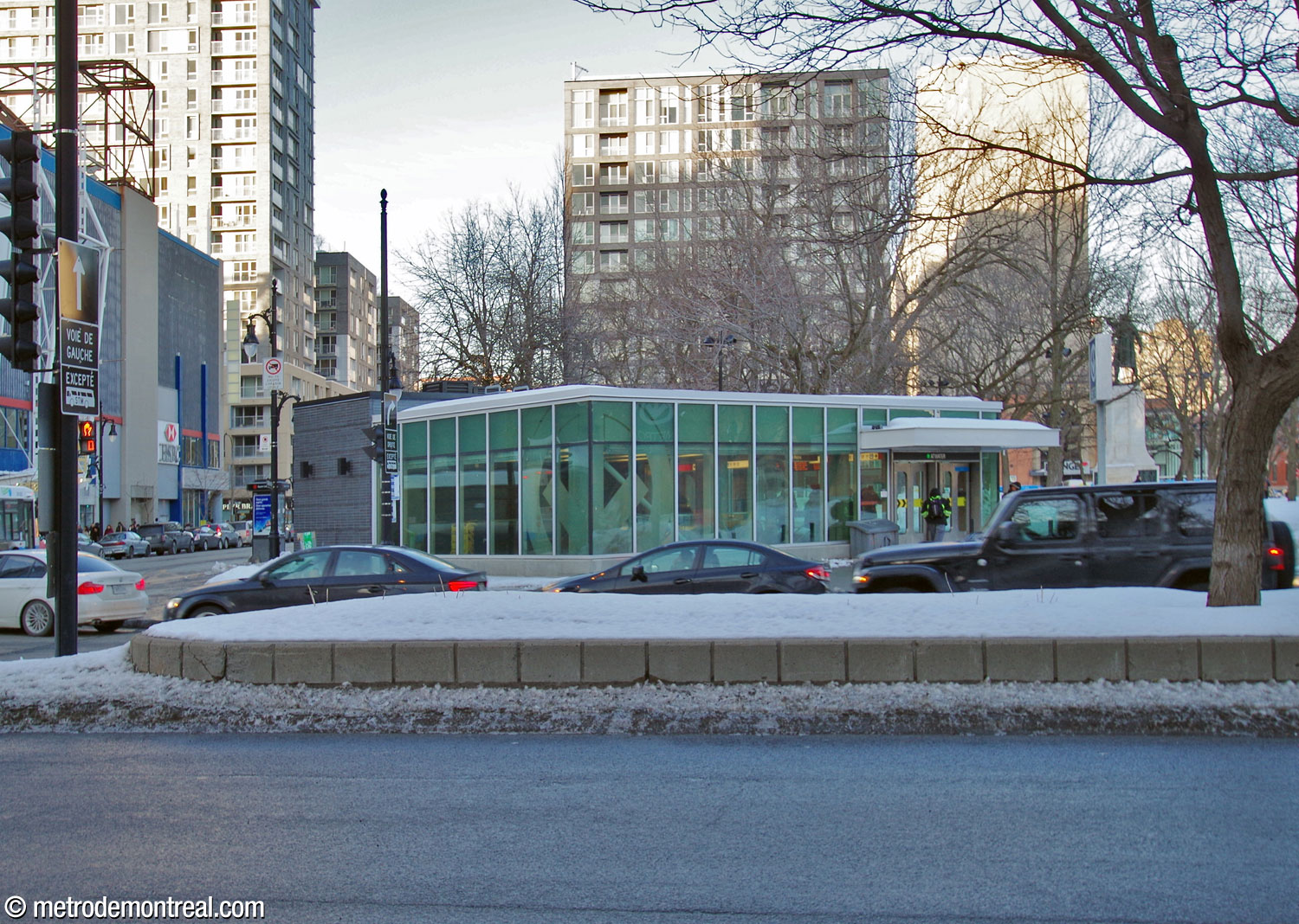 Exterior of the Sainte-Catherine headhouse, seen from across Avenue Atwater. Much of its circumference is glassed in, with a large metro logo etched into the corner windows.