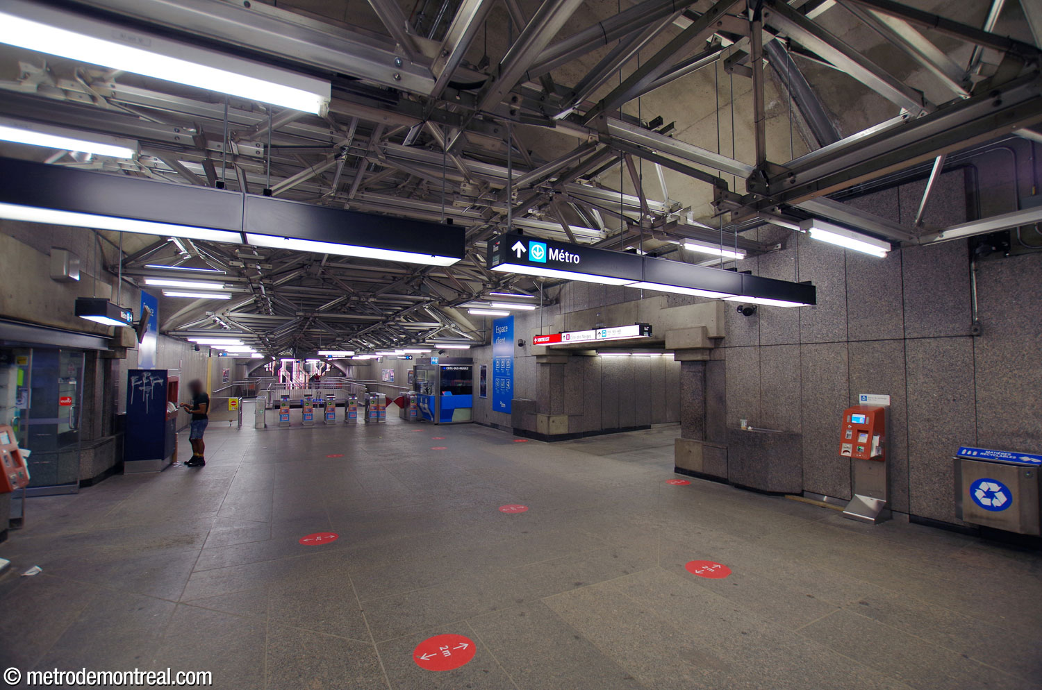 The linear tunnel containing Côte-des-Neiges station's transept, ticket hall, and west access. Next to the ticket booth (right), a perpendicular tunnel crosses under Chemin de la Côte-des-Neiges to reach the east access. Overhead is the signature metal spaceframe that holds the light fixtures and signage.