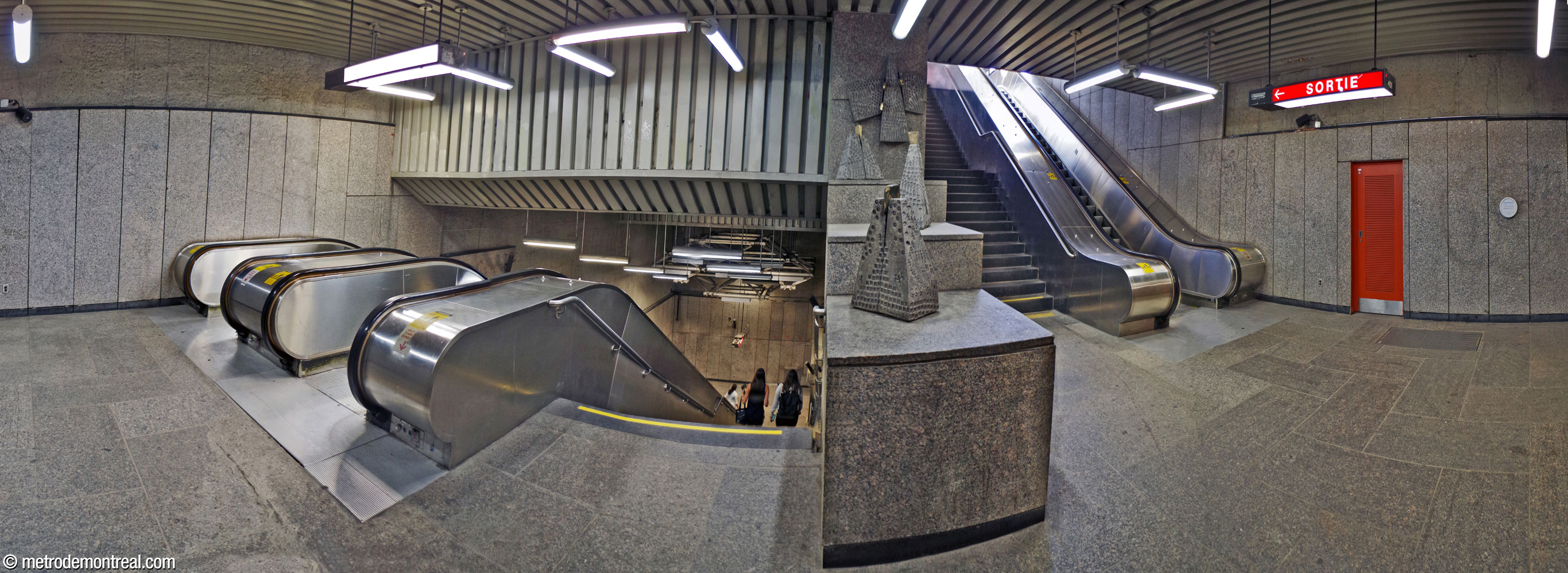 Landing of the stairwell down from the Côte-des-Neiges Est entrance (right). Unusually, the stairs up from the landing are accompanied by only one escalator while the stairs down from it have two. The low wall between the two staircases is home to four aluminum and bronze sculptures by Bernard Chaudron.