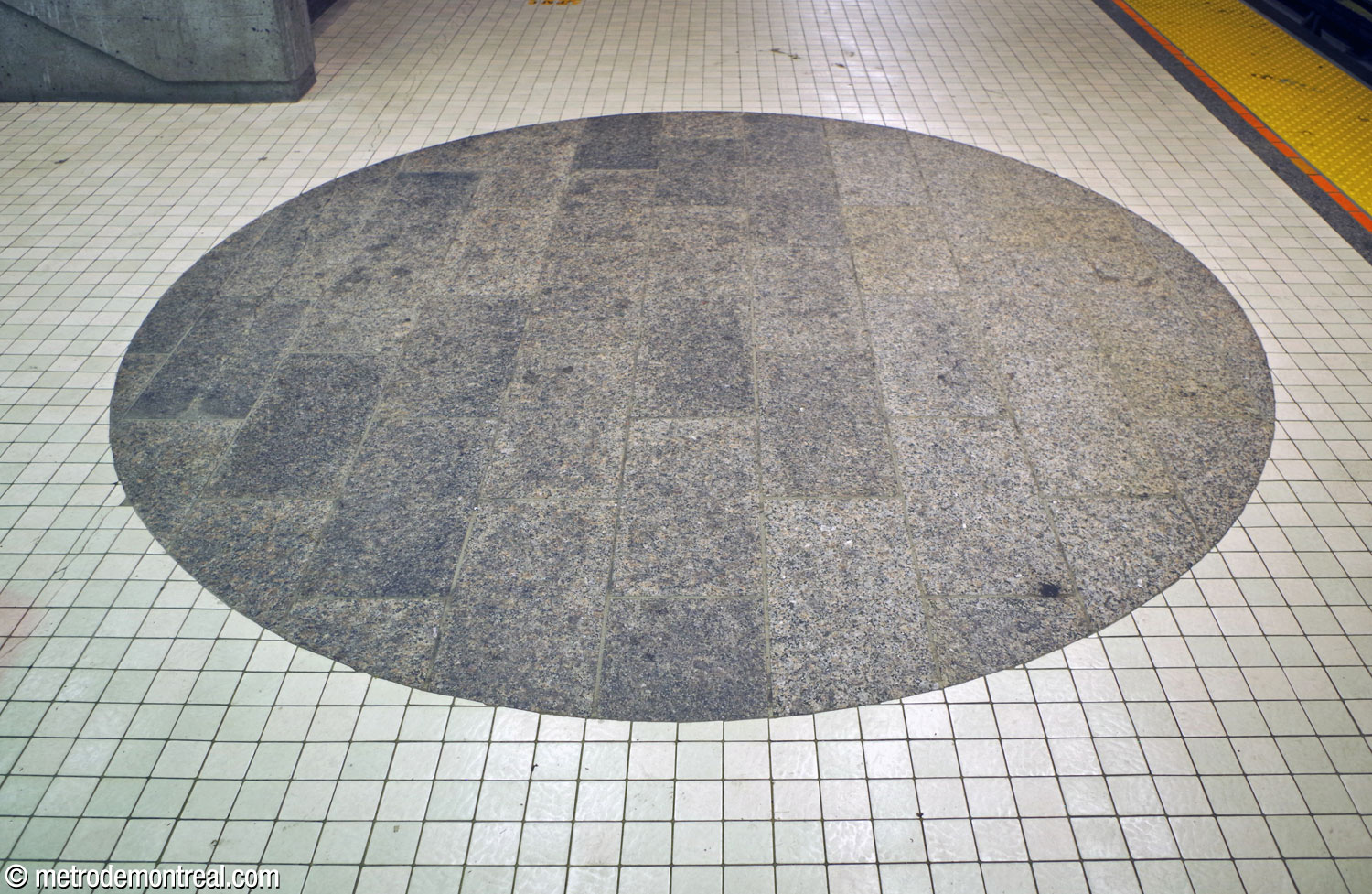 A circle of grey stone slabs punctuates the white floor tiling on the platforms, under the crossover leading to the western access.